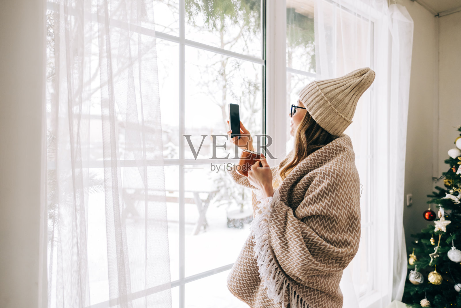 一名年轻的白人妇女站在家里的窗户边，在下雪的早晨拍照。照片摄影图片