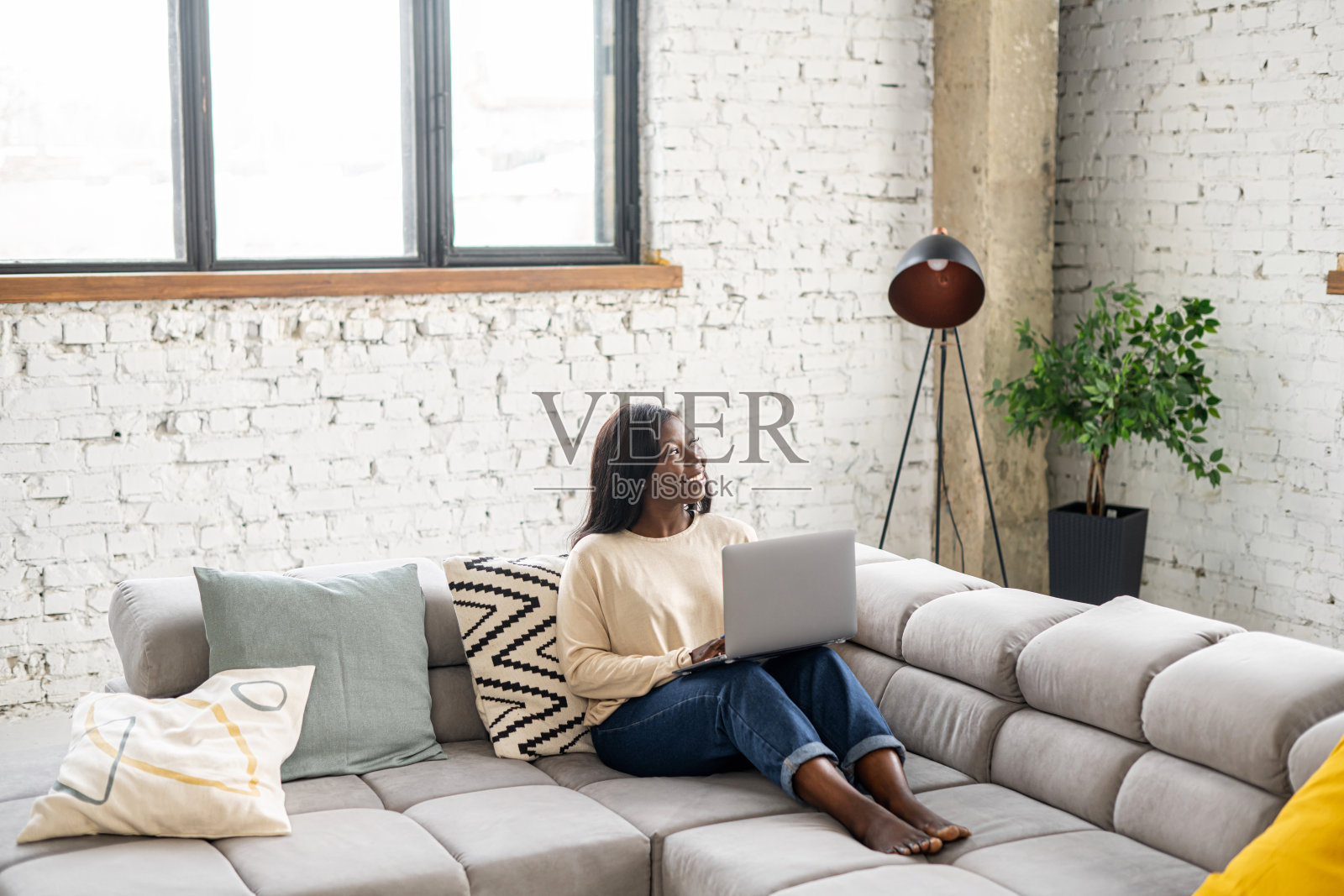 一个多种族的年轻女人在家里玩笔记本电脑。冷静的女孩坐在舒适的沙发上，看着窗前的笔记本电脑屏幕。上网，看电视剧照片摄影图片