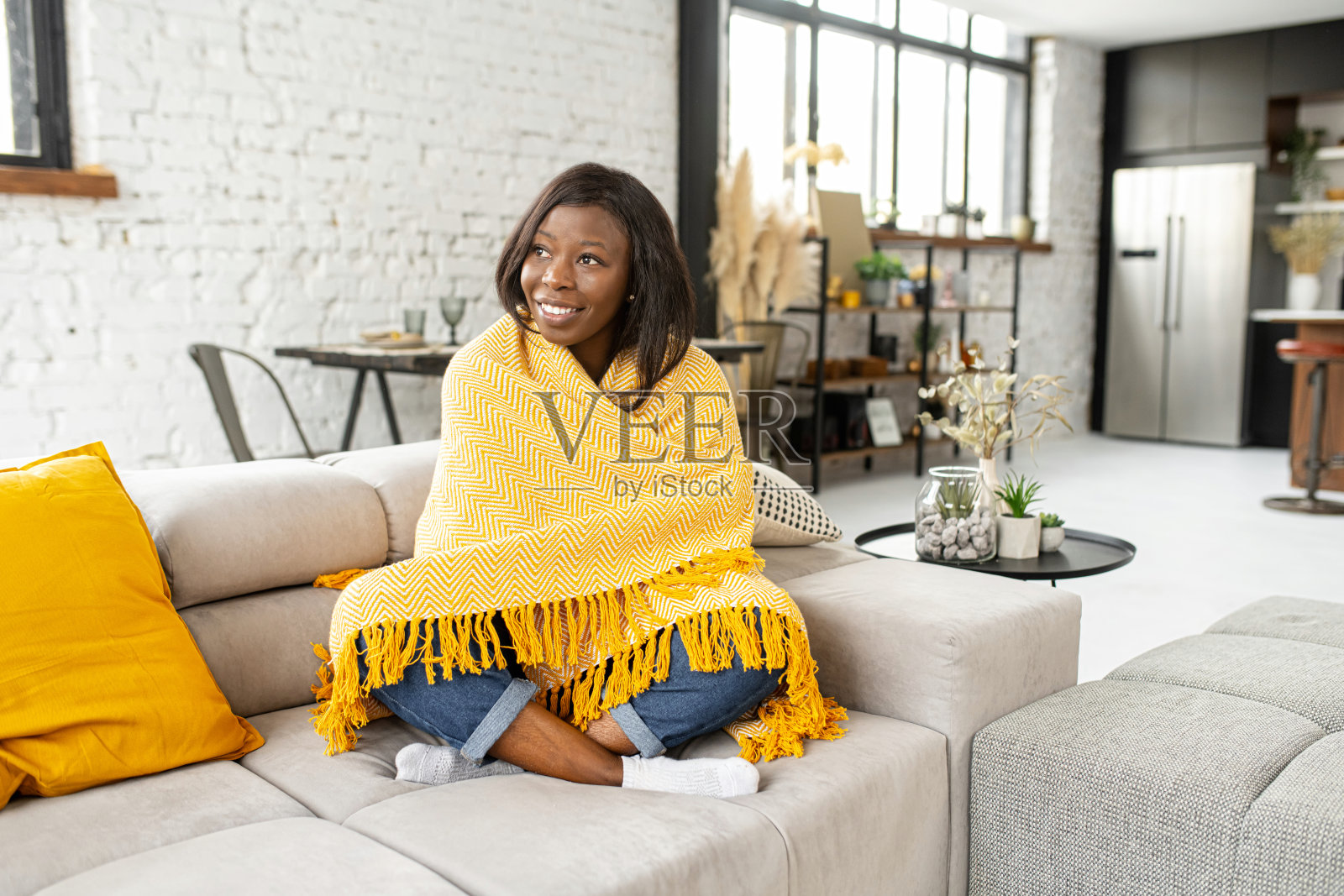 早上好。年轻的多种族少女盖着舒适的黄色柔软的毯子，呆在家里温暖的沙发上，看着窗户照片摄影图片