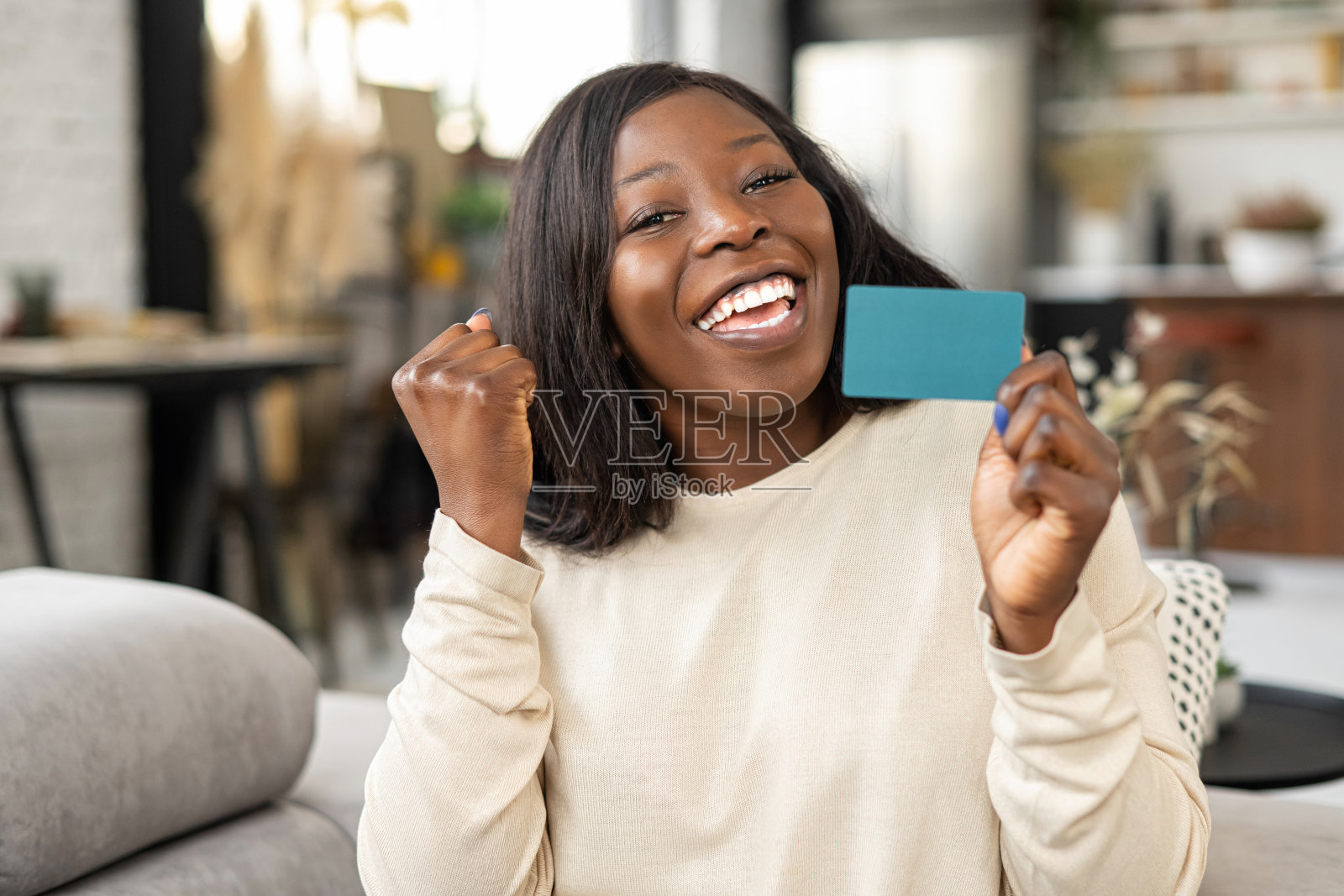 快乐的年轻女子高兴地微笑着，而坐在沙发上的信用卡和看着相机。快乐的混血女孩，拿着信用卡照片摄影图片