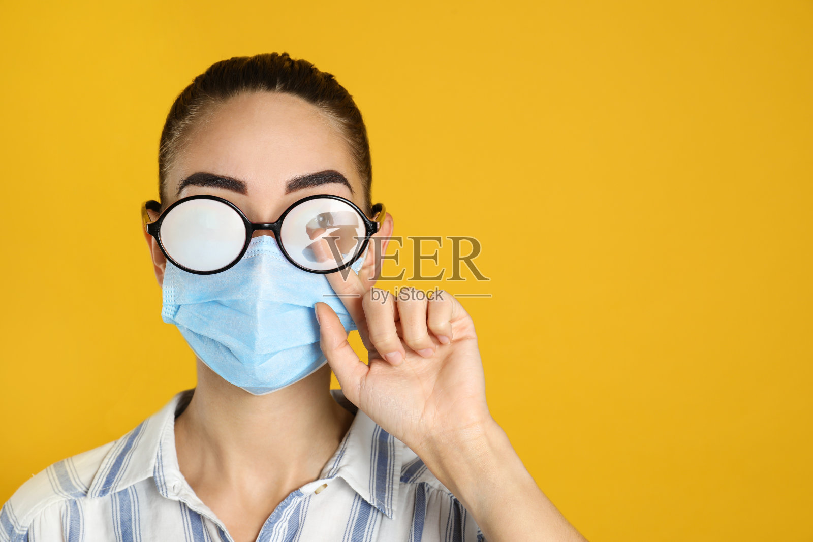 妇女在擦拭黄色背景上戴医用口罩造成的雾状眼镜。空间的文本照片摄影图片