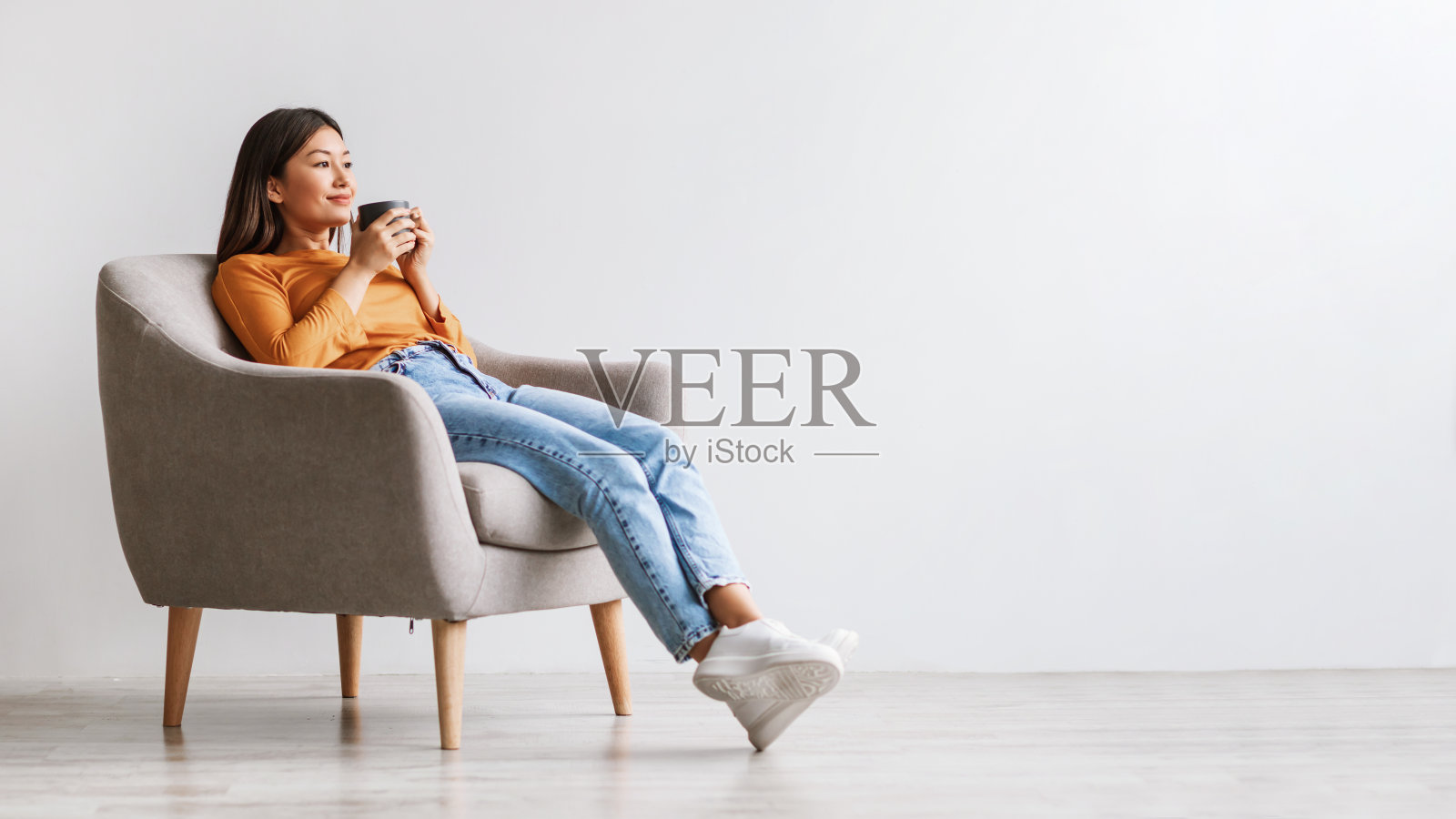 放松的千禧亚洲妇女喝咖啡，休息，在扶手椅靠白墙，自由的空间照片摄影图片