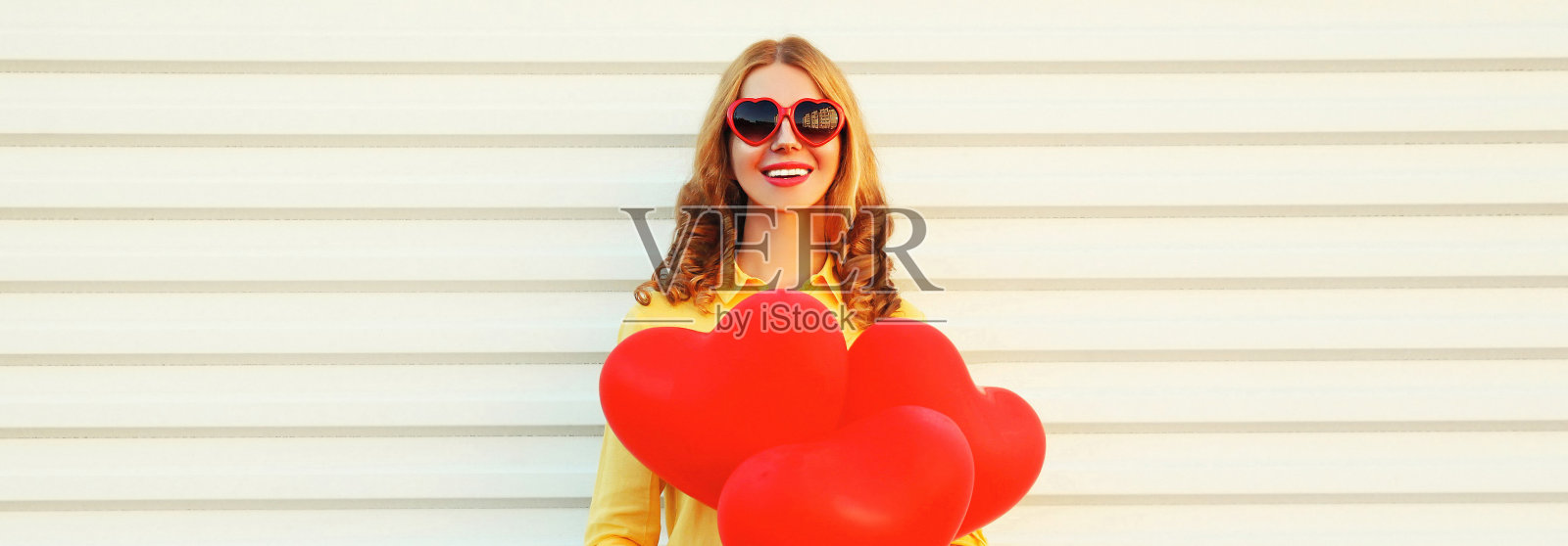 快乐微笑的年轻女子的肖像与一束红色的心形气球穿着黄色的衬衫，在白色的背景太阳镜照片摄影图片