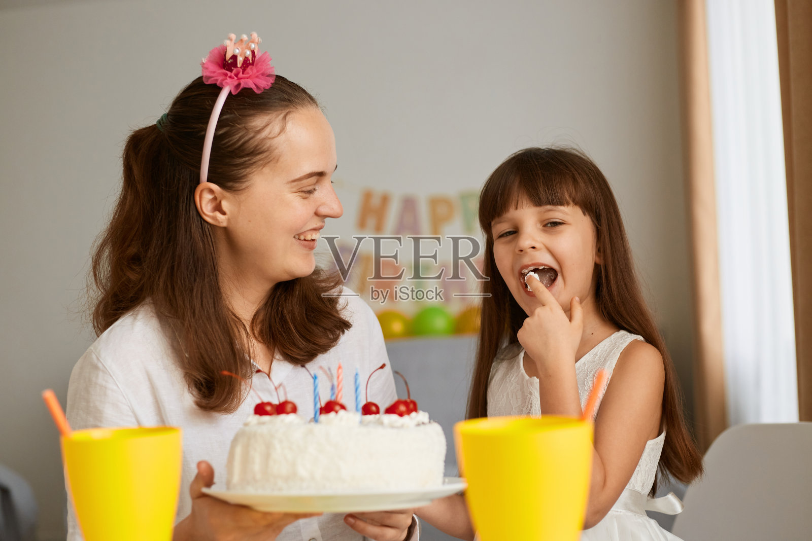 笑脸盈盈的母亲带着女儿庆祝生日，母亲捧着蛋糕，小女孩带着兴奋的表情品尝甜点，背景是节日装饰。照片摄影图片