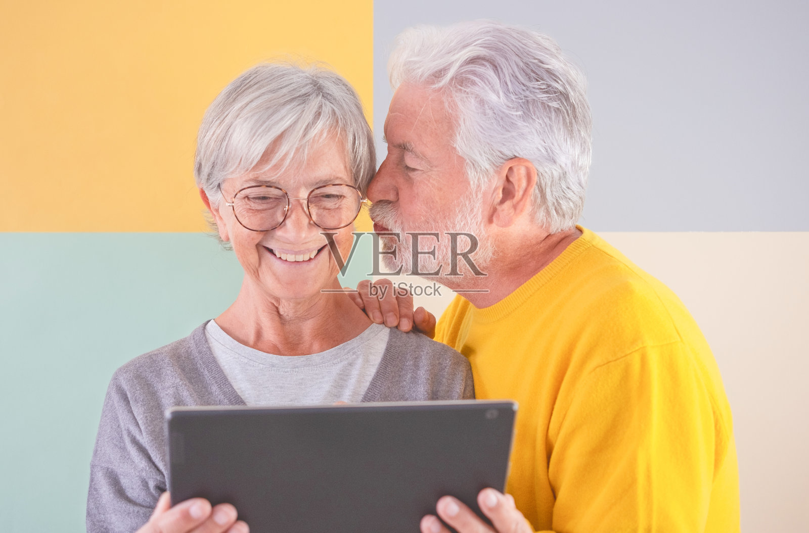 成熟微笑的白人老年夫妇的肖像站在孤立的彩色背景看同一平板。白发老人亲吻他的妻子。退休男女使用科技和社交媒体照片摄影图片