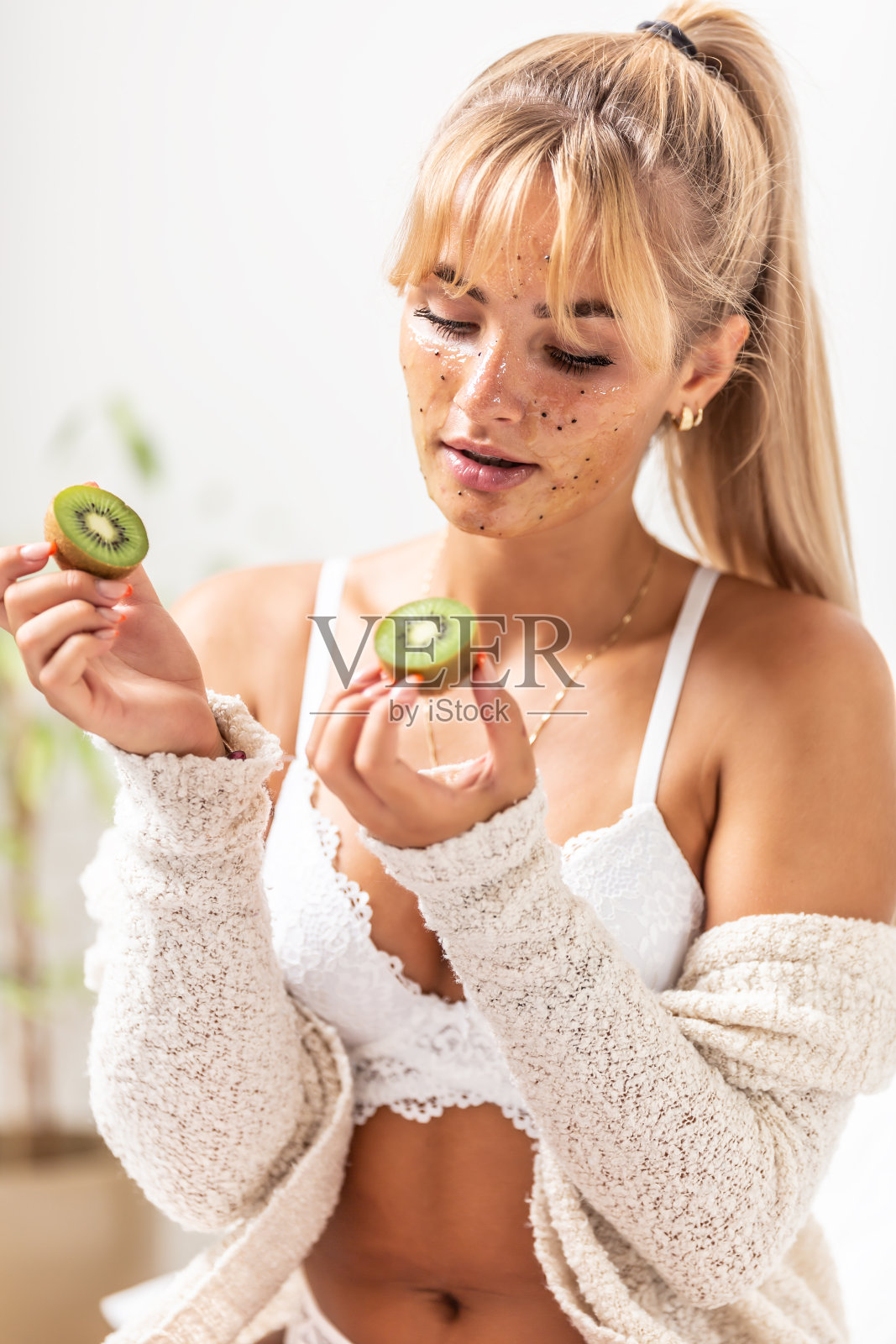 美丽的年轻金发女孩坐在白色内衣床上应用猕猴桃脸磨砂在她的脸，手握新鲜的猕猴桃。照片摄影图片
