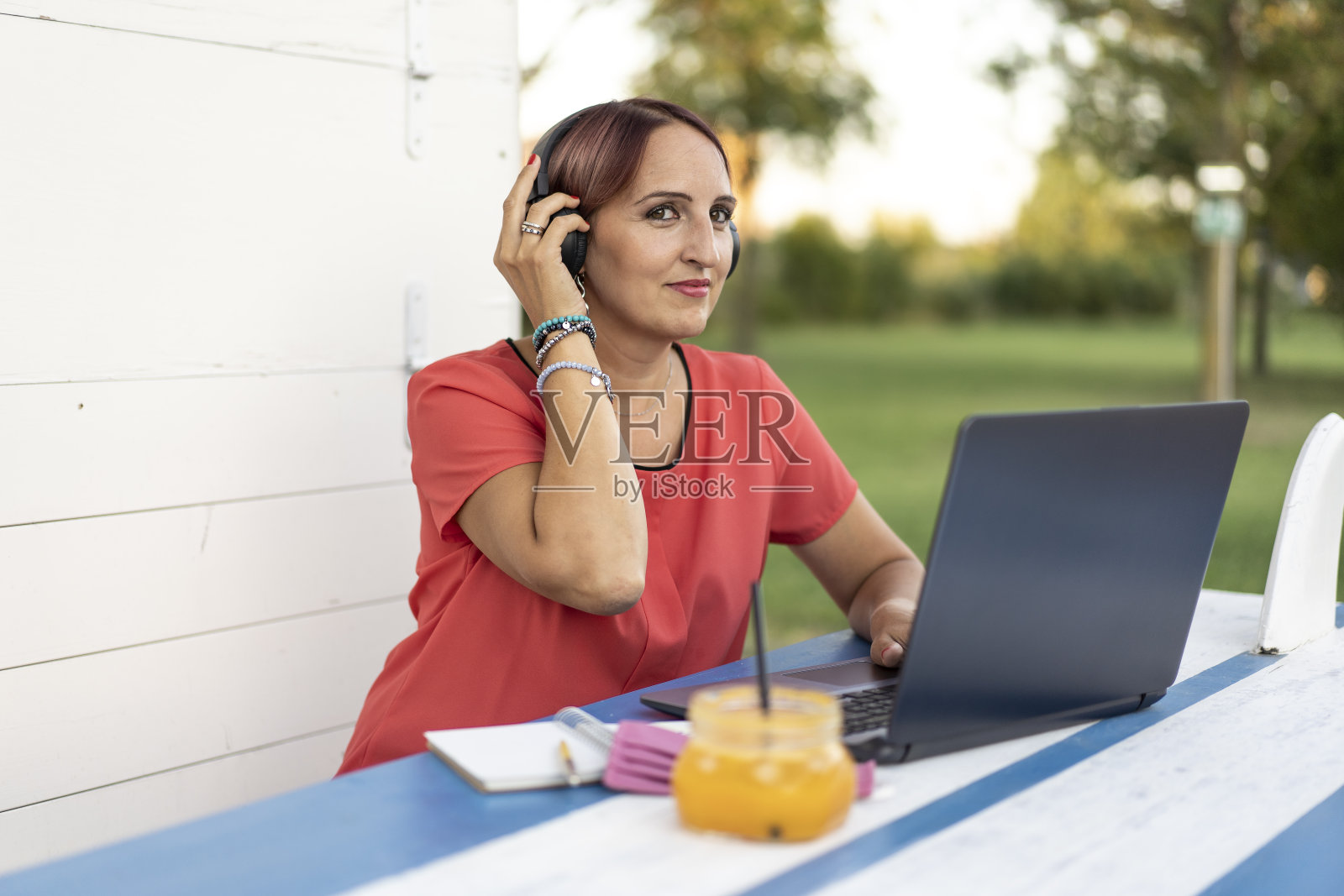 在公园网上工作的女商人。自由职业者使用技术在任何地方工作。一名女子在户外用耳机和笔记本电脑与顾客交谈。照片摄影图片