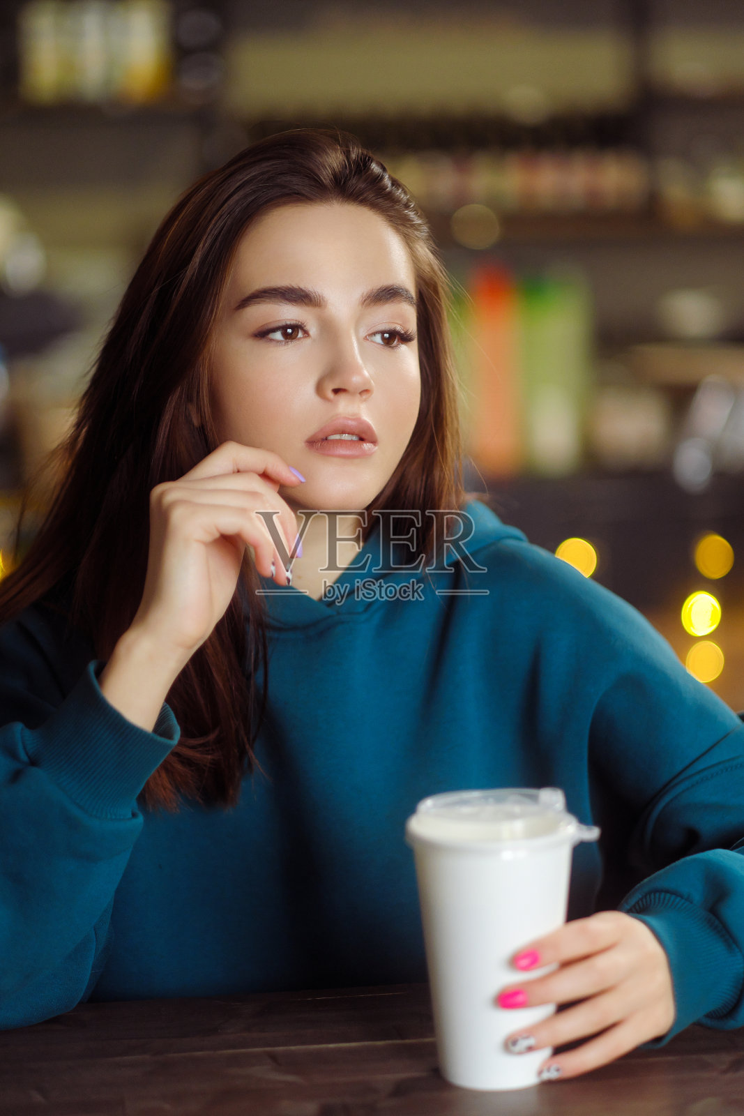 年轻白人黑发女人的肖像，在绿色帽衫，坐在咖啡馆与纸杯。照片摄影图片