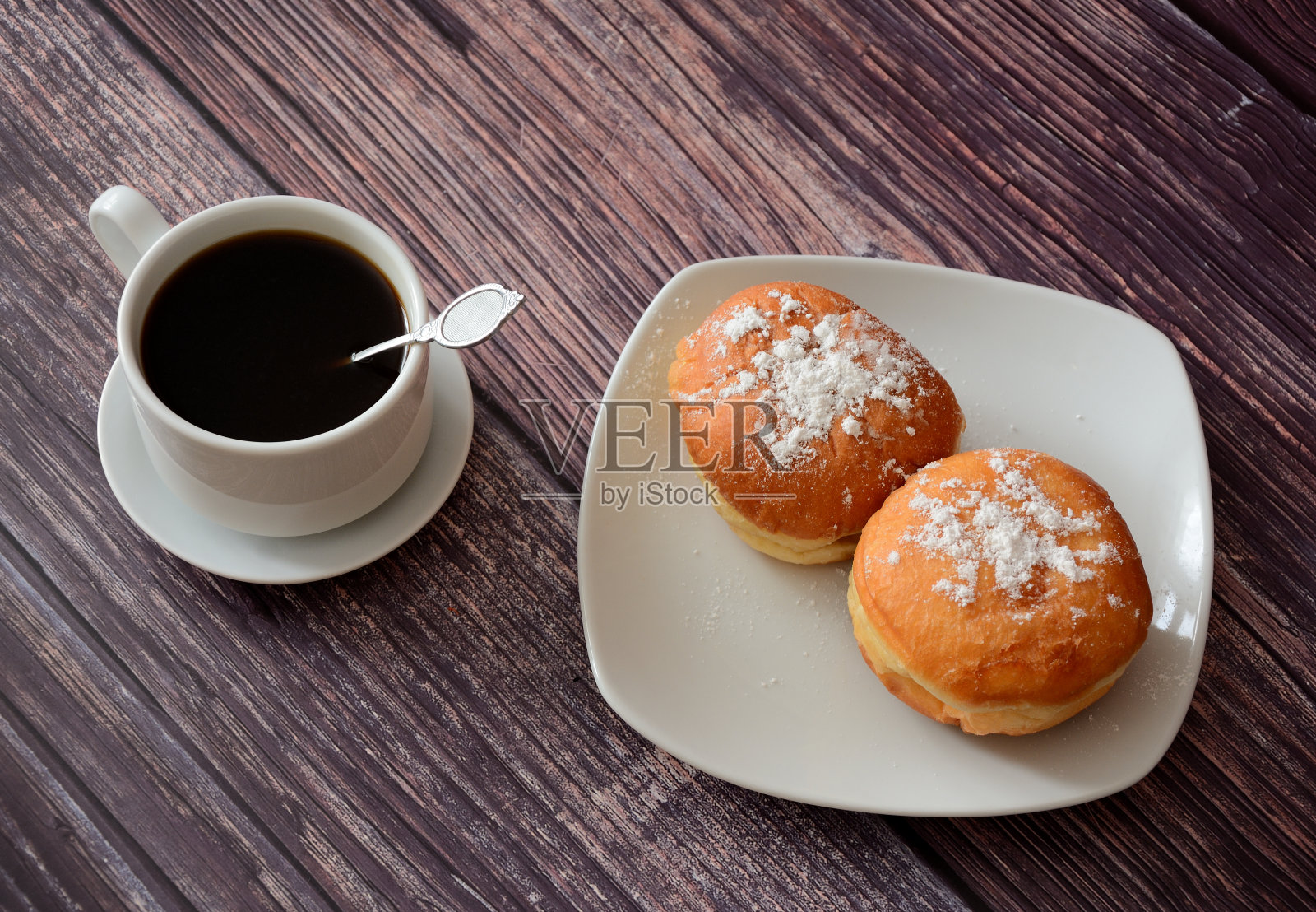 木桌上有一杯新鲜的黑咖啡和一个装有两个糖粉甜甜圈的盘子。照片摄影图片