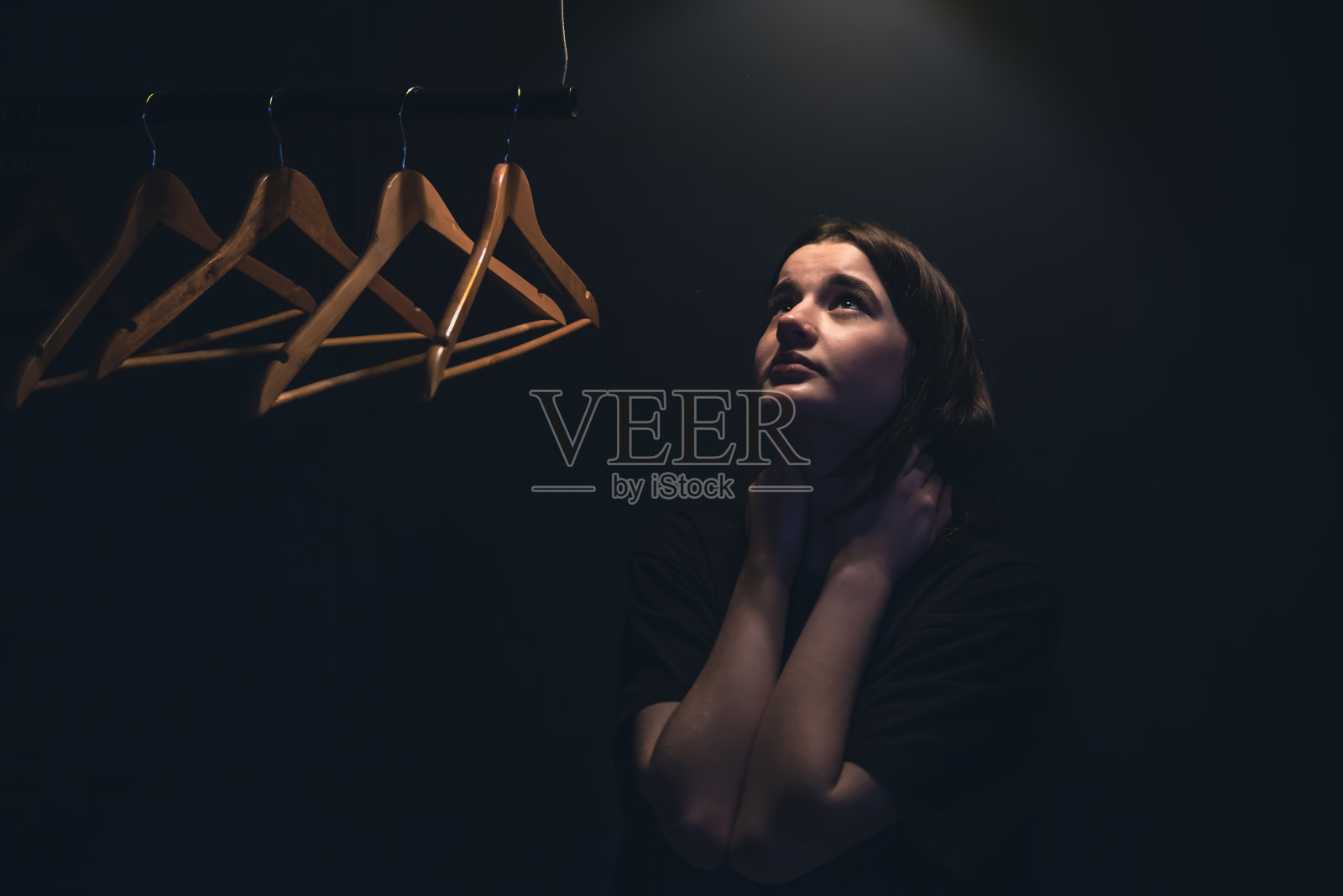 一个年轻的女人在黑暗中，在一盏灯下靠近空衣架。照片摄影图片