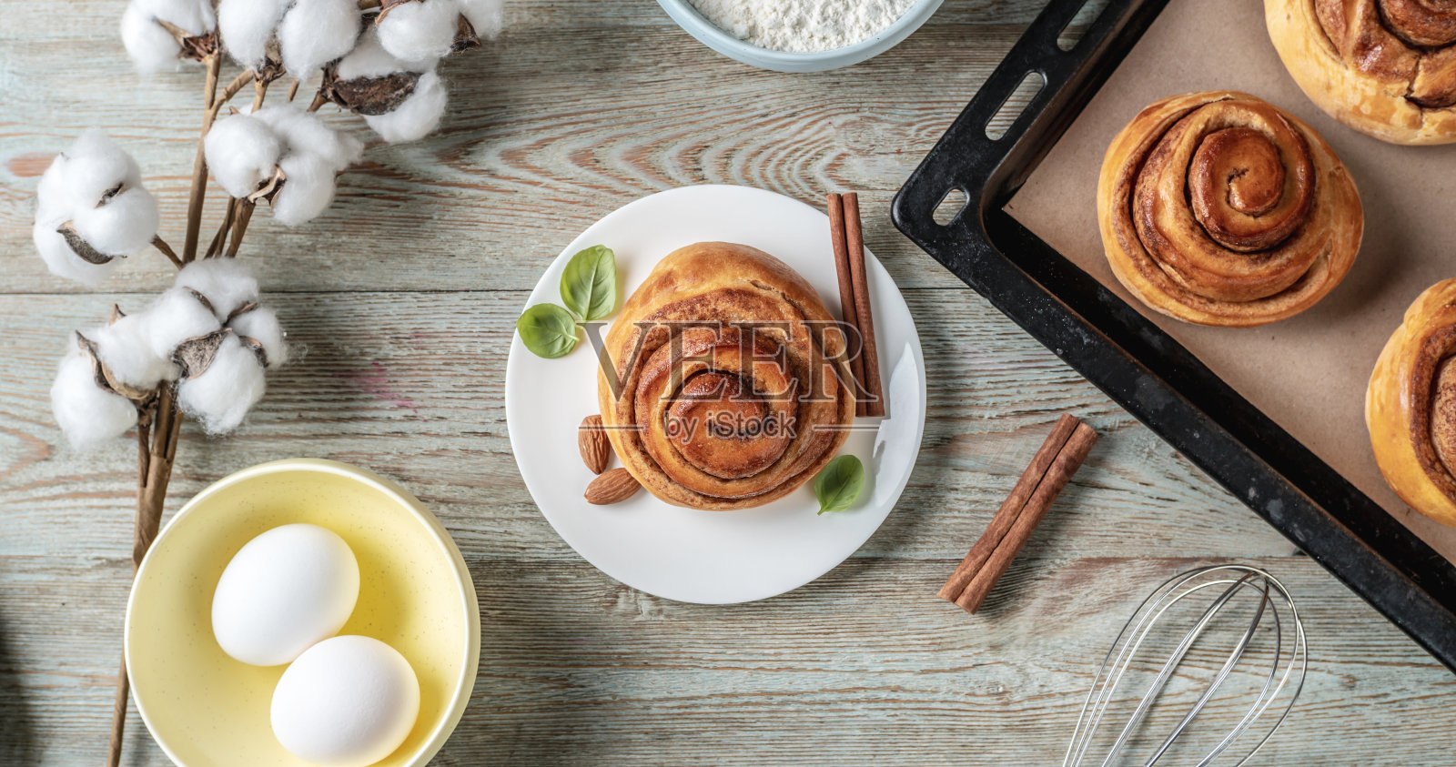 桌子上有一个烤盘，上面放着新鲜的肉桂卷、烹饪材料和开胃的小圆面包。美味的自制糕点概念和舒适的氛围。俯视图照片摄影图片
