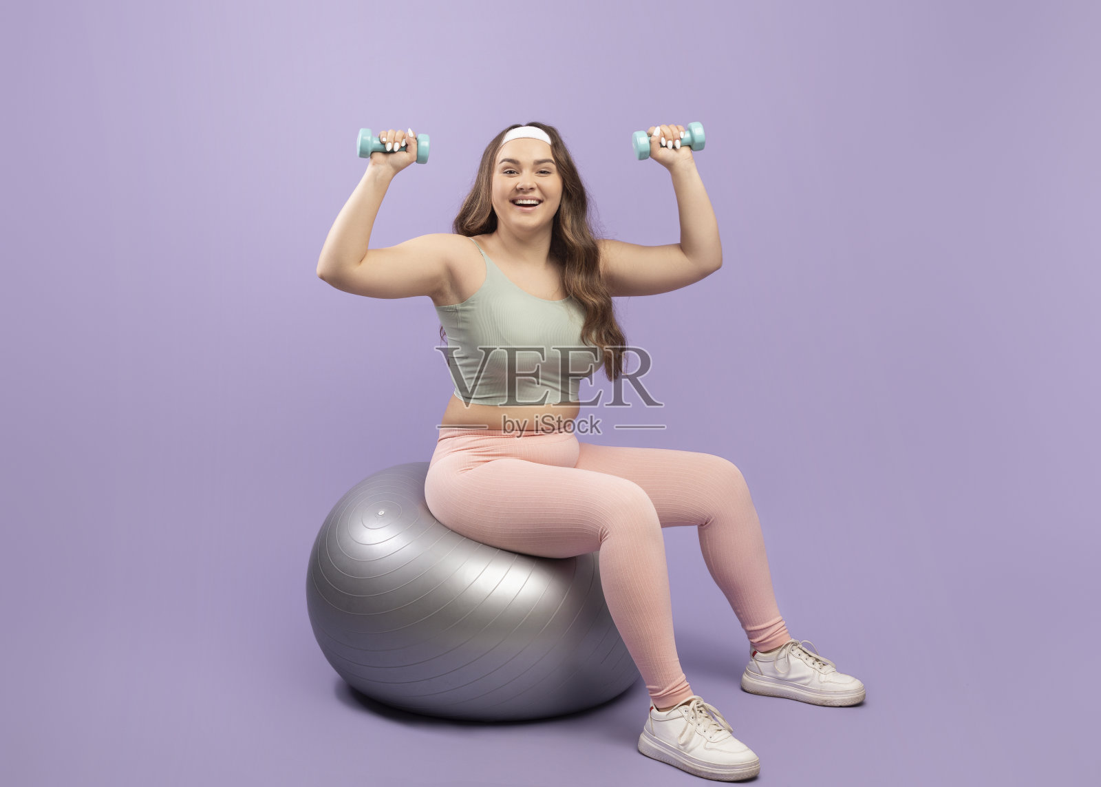 年轻的欧洲胖女人，穿着运动服，在健身球上举哑铃照片摄影图片