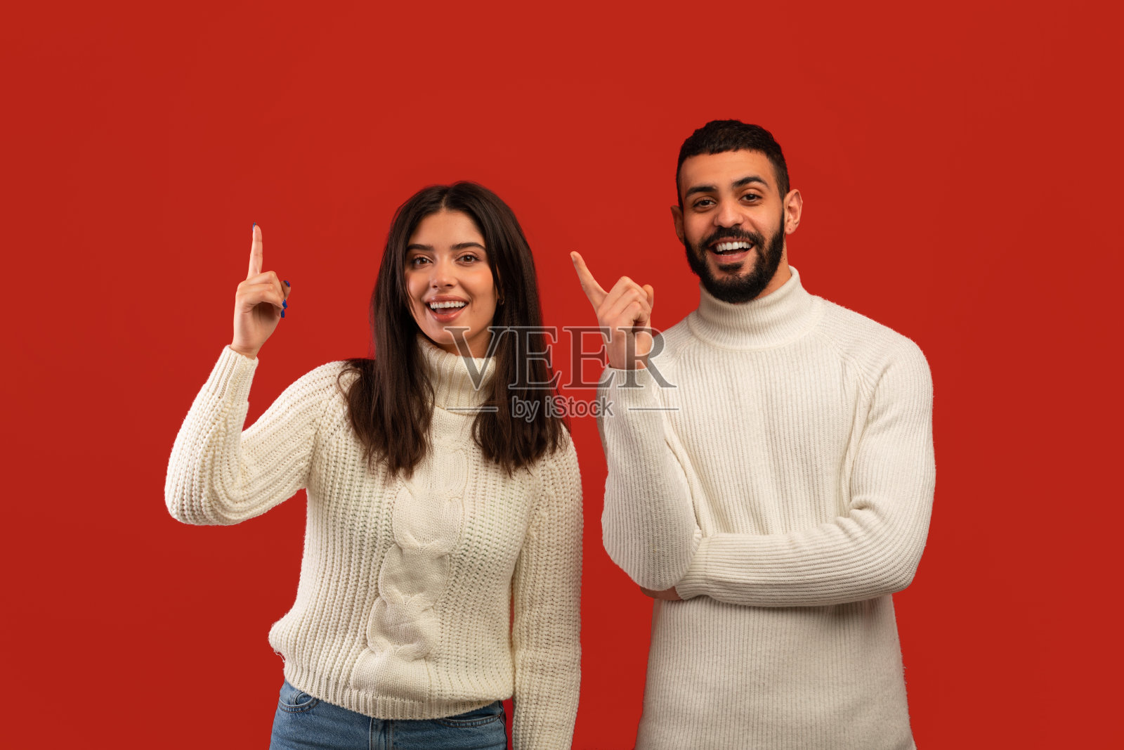 一对中东夫妇站在红色的演播室背景上，指着手指，对着镜头微笑，有很棒的想法照片摄影图片