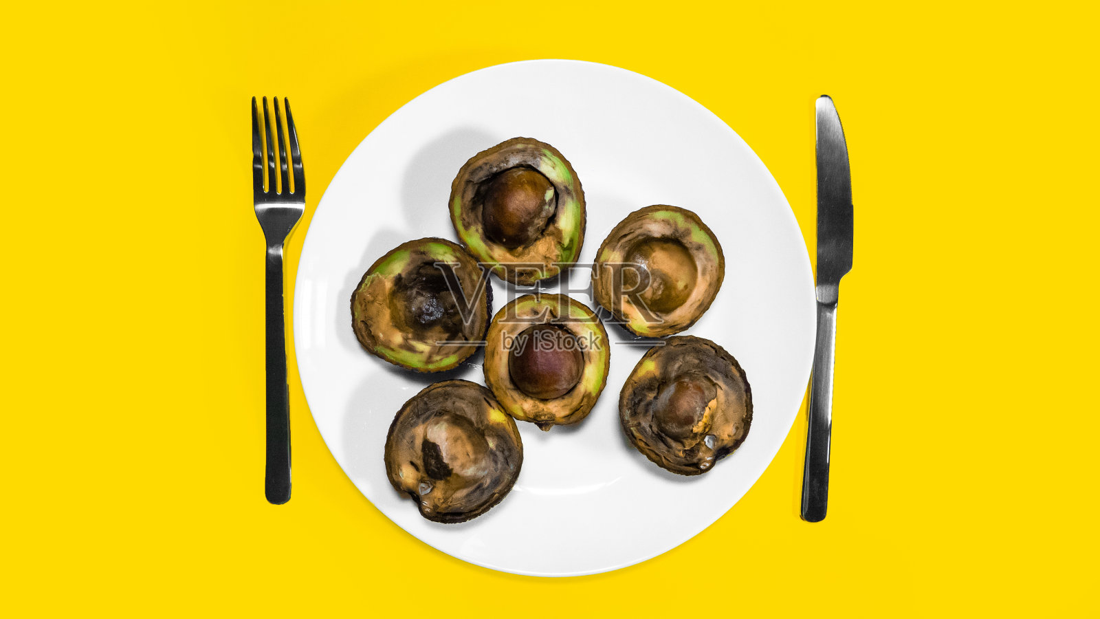 坏鳄梨的一半放在盘子上与叉和刀黄色背景。照片摄影图片