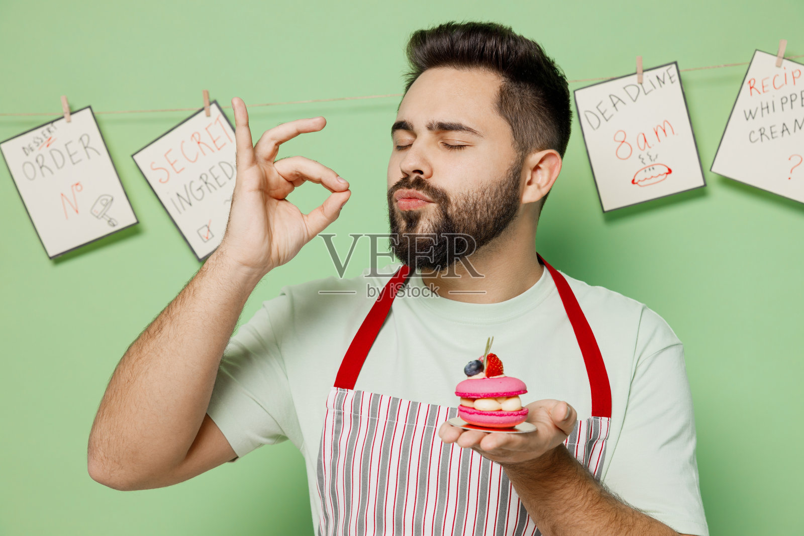年轻的男性厨师，糖果师，面包师，穿着条纹围裙，拿着粉红色的小蛋糕松饼马卡龙，展示belissimo完美的姿态，孤立在素色的淡绿色背景烹饪食物的概念。照片摄影图片