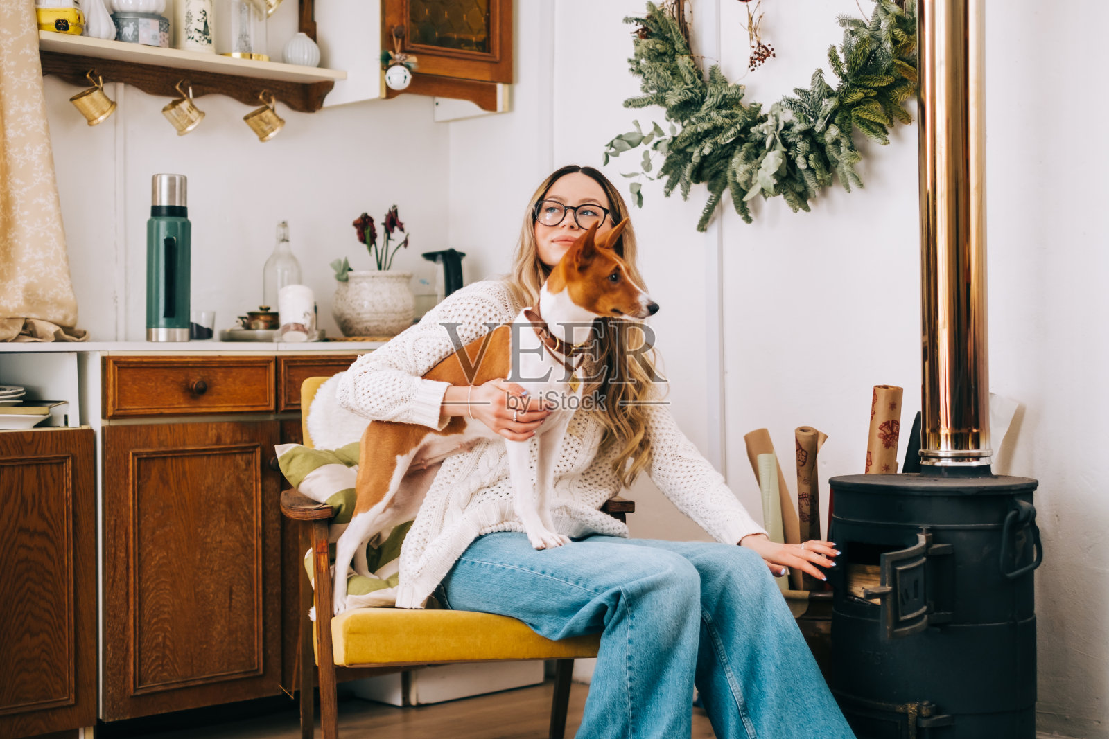 年轻的白人妇女坐在扶手椅附近的壁炉和她的狗在冬季假期。照片摄影图片