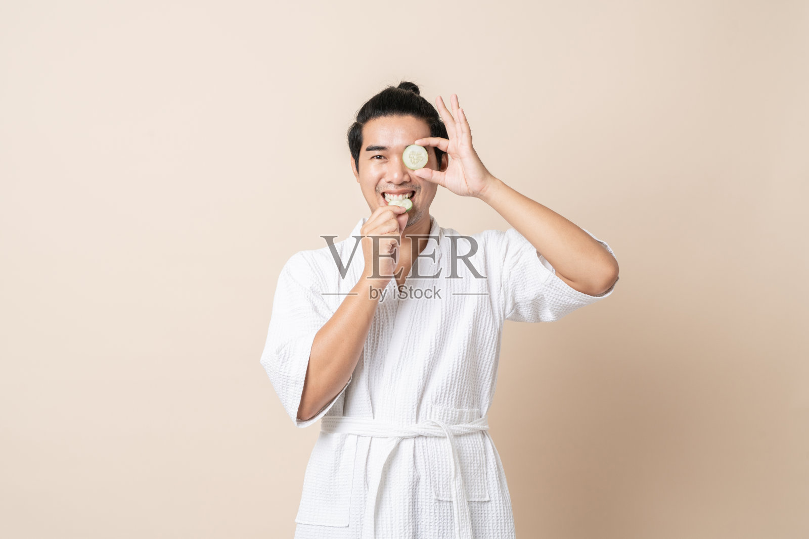 亚洲男人开心的微笑着穿着浴袍或水疗服，拿着黄瓜片做面膜。照片摄影图片