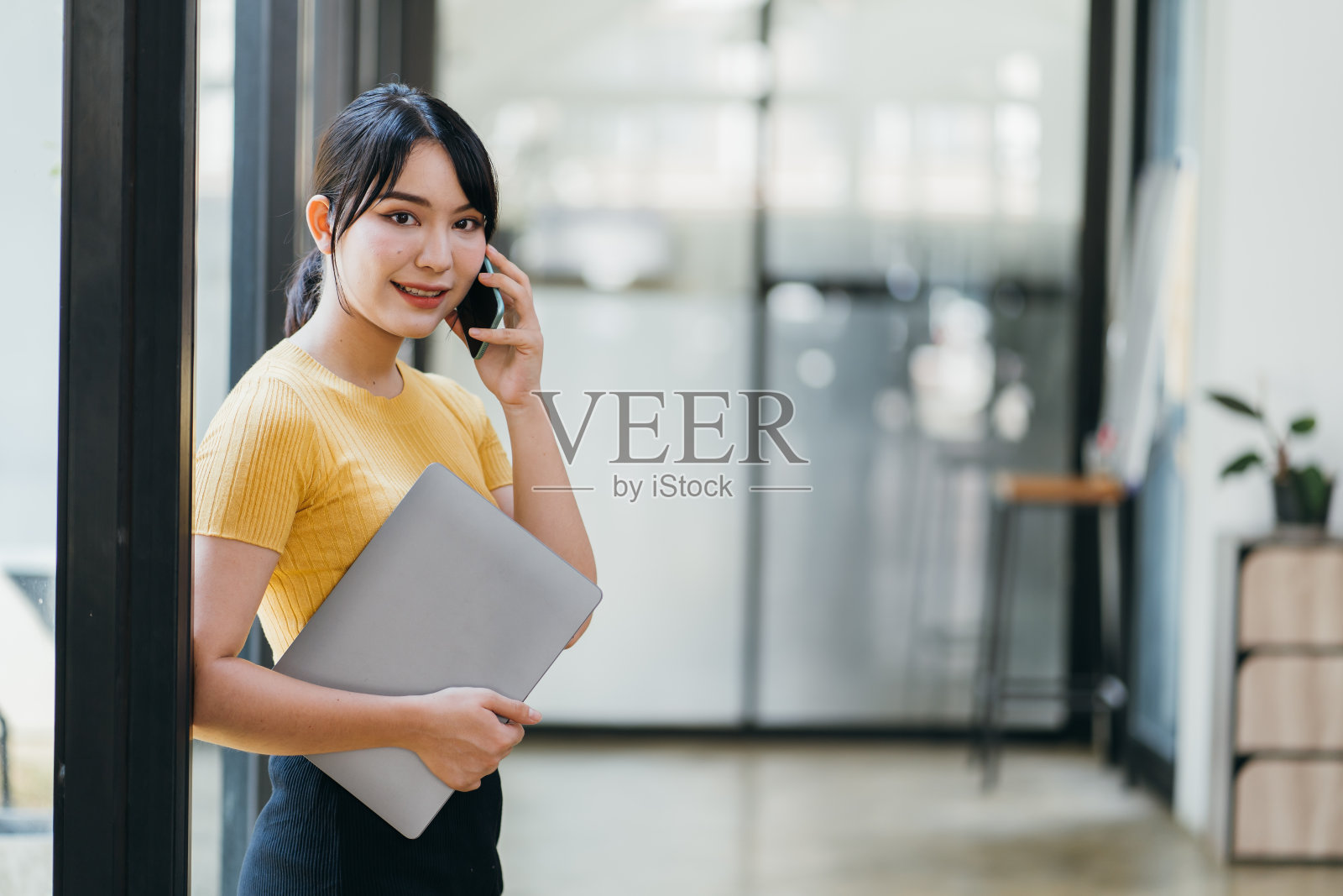 侧面的亚洲女性穿着正式，微笑，看着相机，在现代办公室浏览智能手机。照片摄影图片