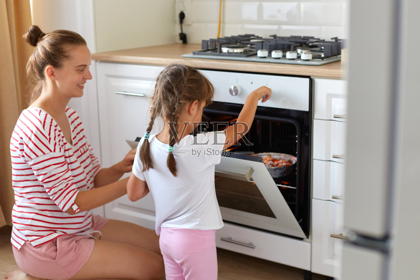快乐的年轻女子和可爱的女儿穿着休闲服装在厨房烤甜点，教孩子做纸杯蛋糕，坐在烤炉旁的地板上，在家做饭。照片摄影图片