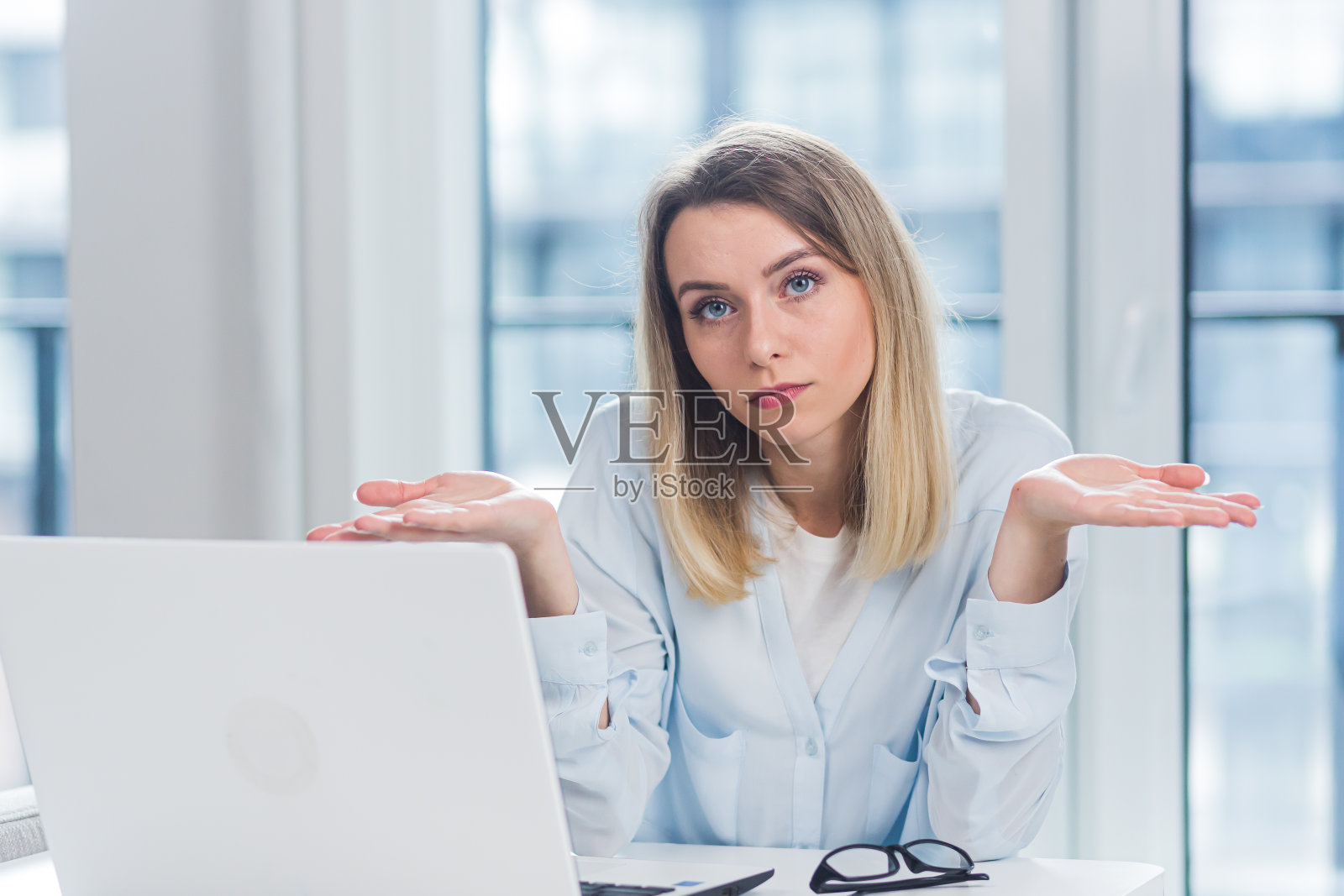 一个年轻的成年金发女商人的特写肖像看着在办公室或在蓝色窗户背景的室内的相机。在室内穿着正式服装面对自信的女经理照片摄影图片