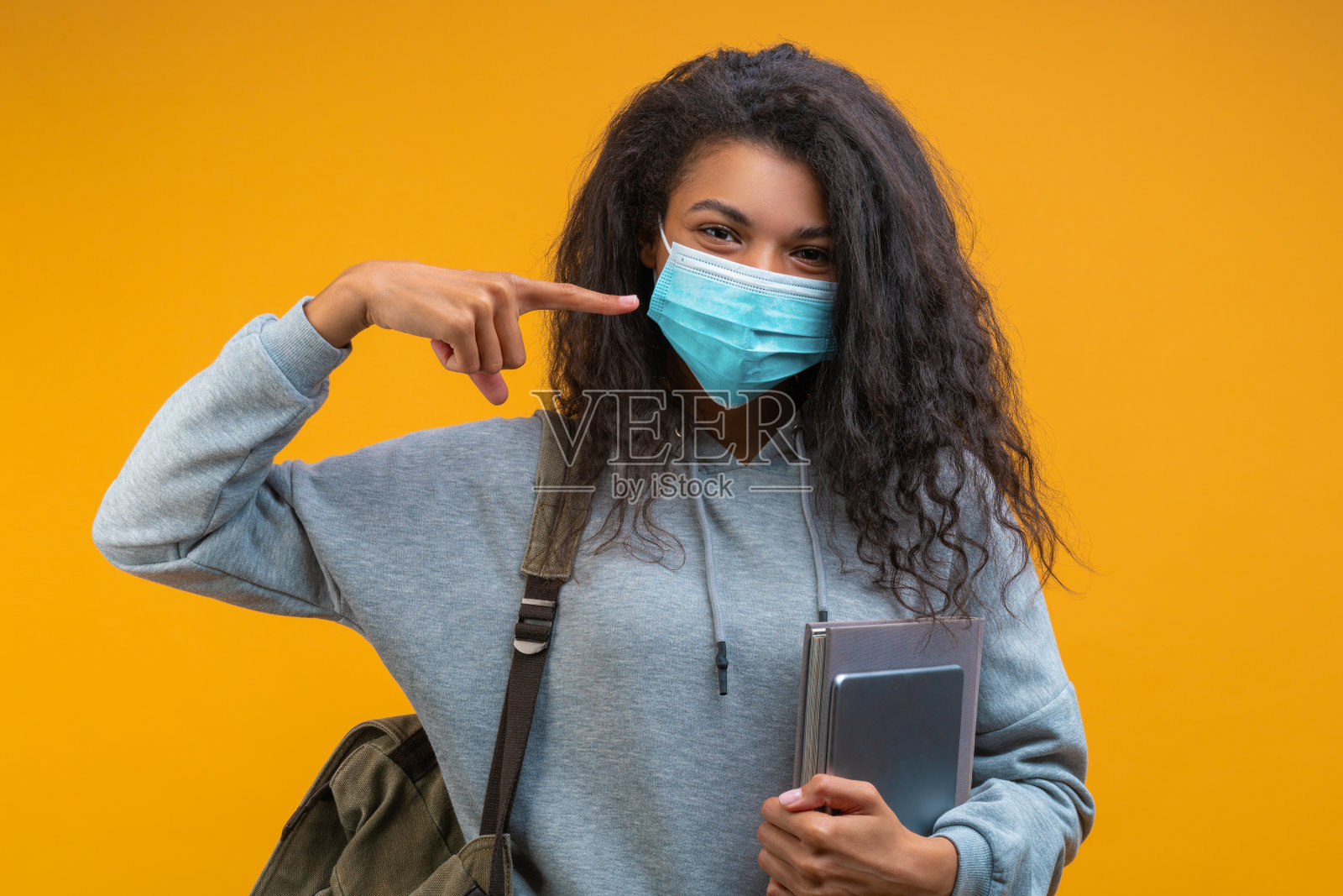 摄影室的肖像，穿着随意的非裔美国女学生背着背包，拿着书和平板电脑，用食指指着她脸上的保护性医疗口罩照片摄影图片