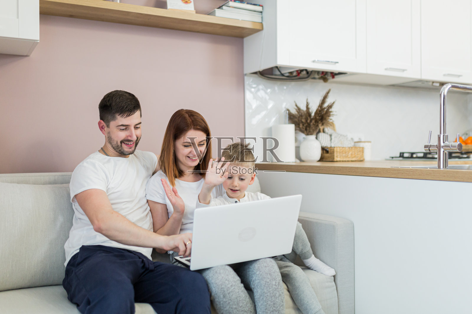 年轻的一家人，妈妈，爸爸，两个孩子，坐在家里房间的沙发上，用笔记本电脑打视频电话，问候，微笑照片摄影图片