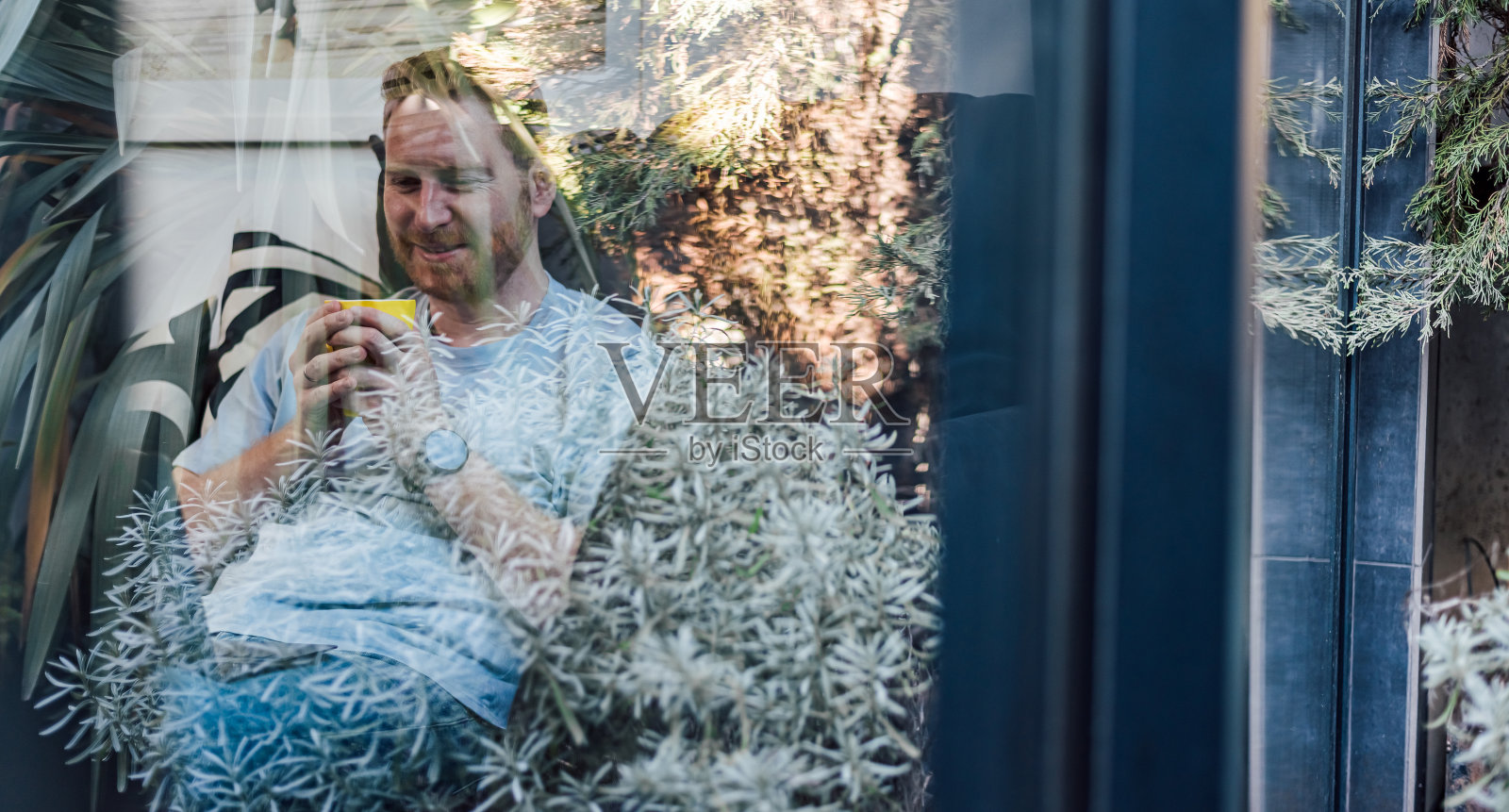 从花园望向窗外，看到一名笑容满面的年轻男子舒舒服服地坐在家里的大沙发上，喝着咖啡或茶，度过闲暇时光照片摄影图片