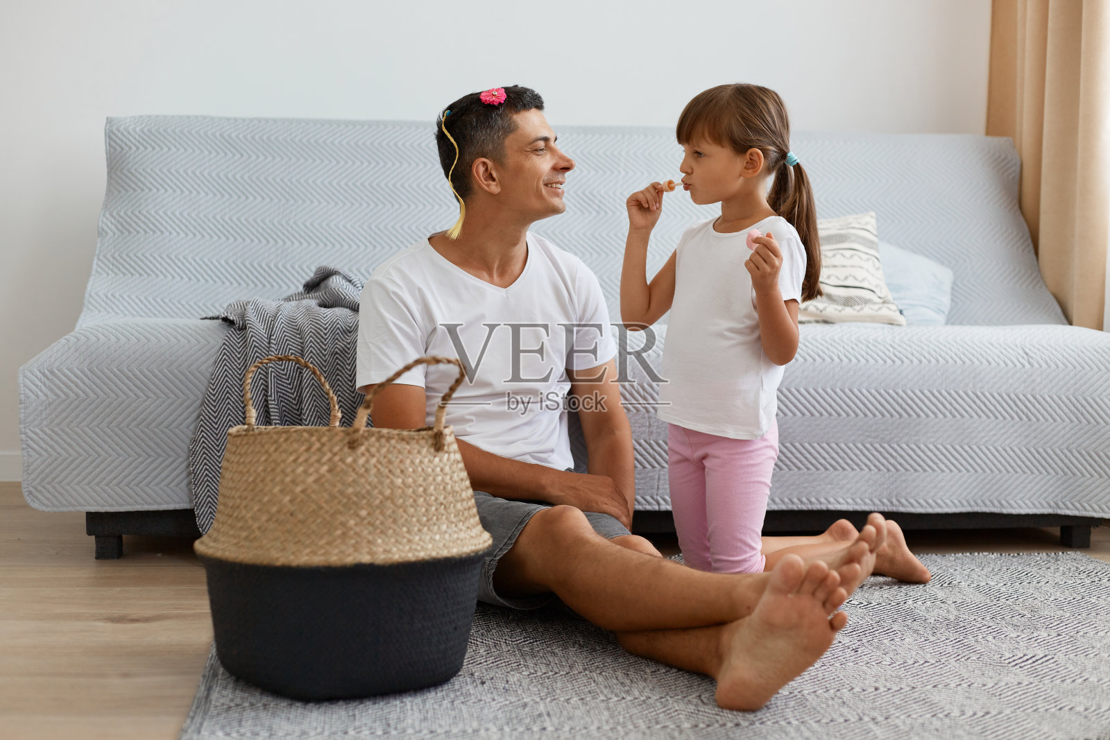 室内拍摄的迷人的白人男子穿着白色休闲风格的t恤和牛仔裤短裤坐在地板上的黑色头发的小女孩，女儿用妈妈的润发油，花时间与爸爸。照片摄影图片