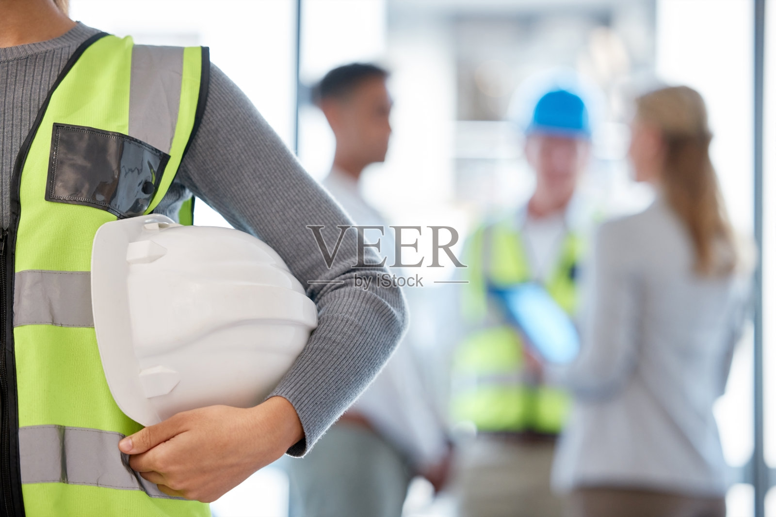 剪裁的镜头，一个不认识的女建筑工人站在她的胳膊和她的同事在背景下的安全帽照片摄影图片