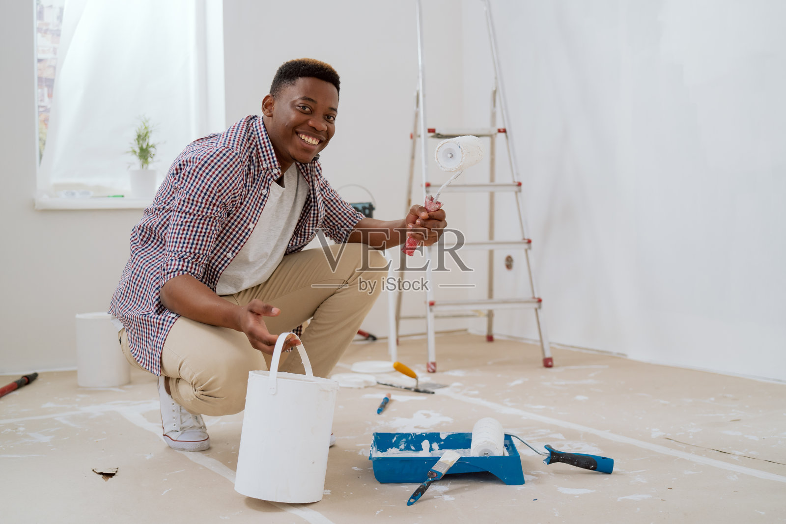 一个穿着棕色牛仔裤和红色格子衬衫、面带微笑的小男孩拿着一个滚筒，拿起一罐白漆。他正在装修一套公寓，正准备粉刷墙壁照片摄影图片