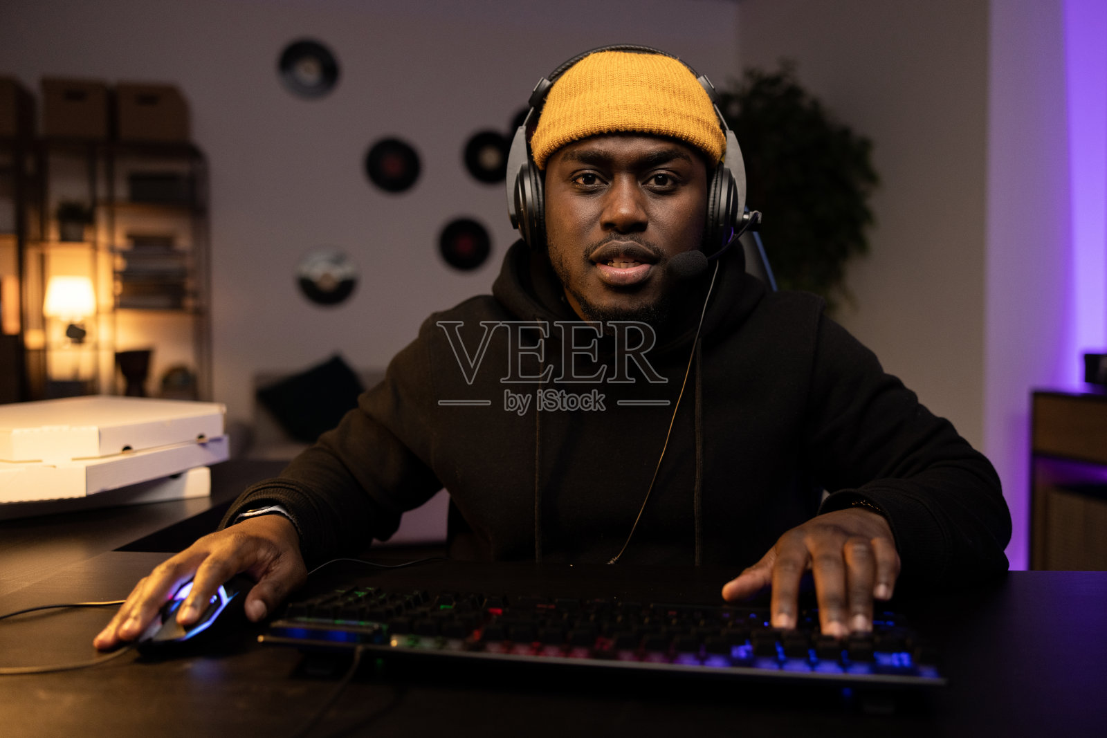 一名戴着黄色帽子戴着耳机的严肃男子正在电脑上认真地玩视频射击游戏。在线多人游戏锦标赛。背景是一个夜晚漆黑的房间，里面堆满了披萨盒照片摄影图片