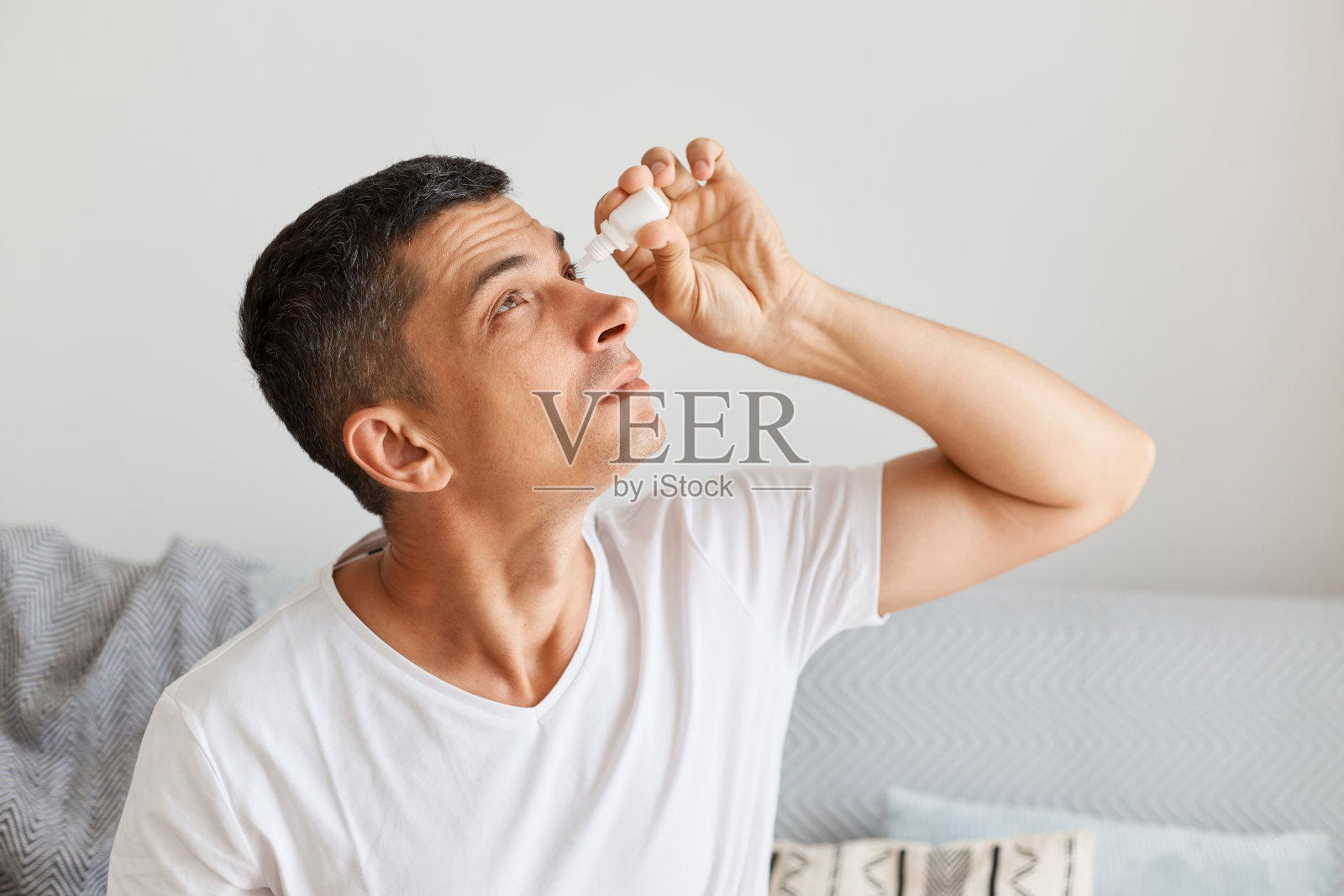 室内拍摄的黑发白人男子使用眼药水，有视力问题，治疗，过度工作的男性使用眼药水治疗眼睛。照片摄影图片