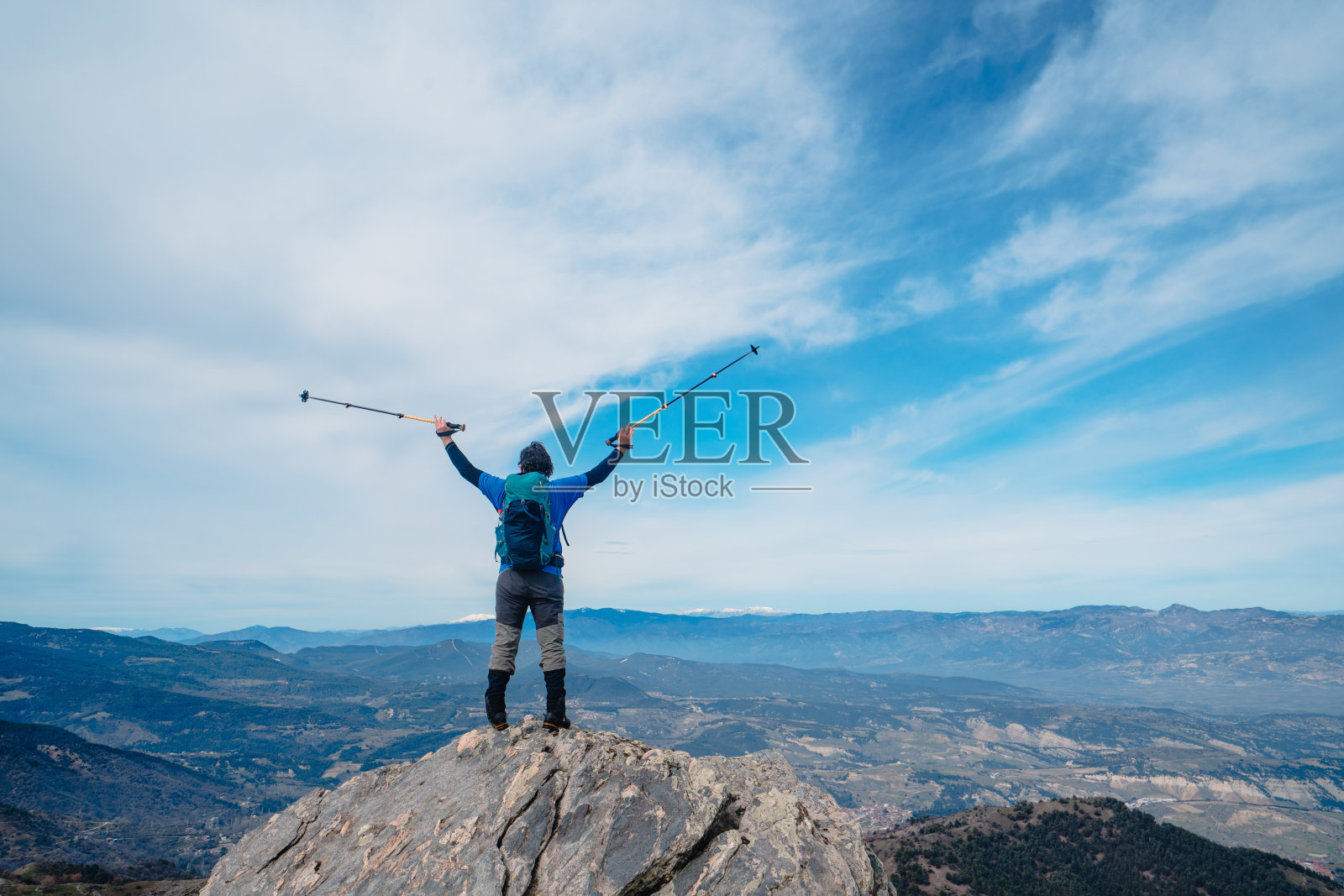 后视图的摄影师登山运动员举起他的手臂到空中，而持有登山杆在一个岩石山的顶峰。照片摄影图片