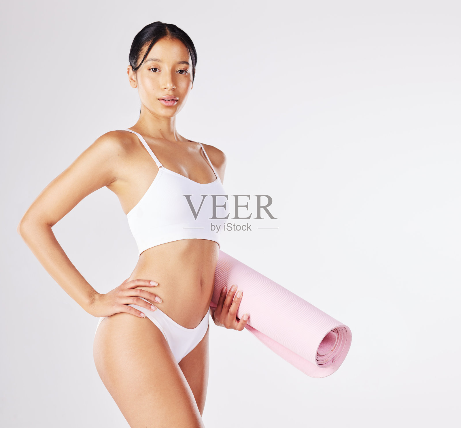 一个健康的年轻女子拿着瑜伽垫，站在工作室的背景下照片摄影图片