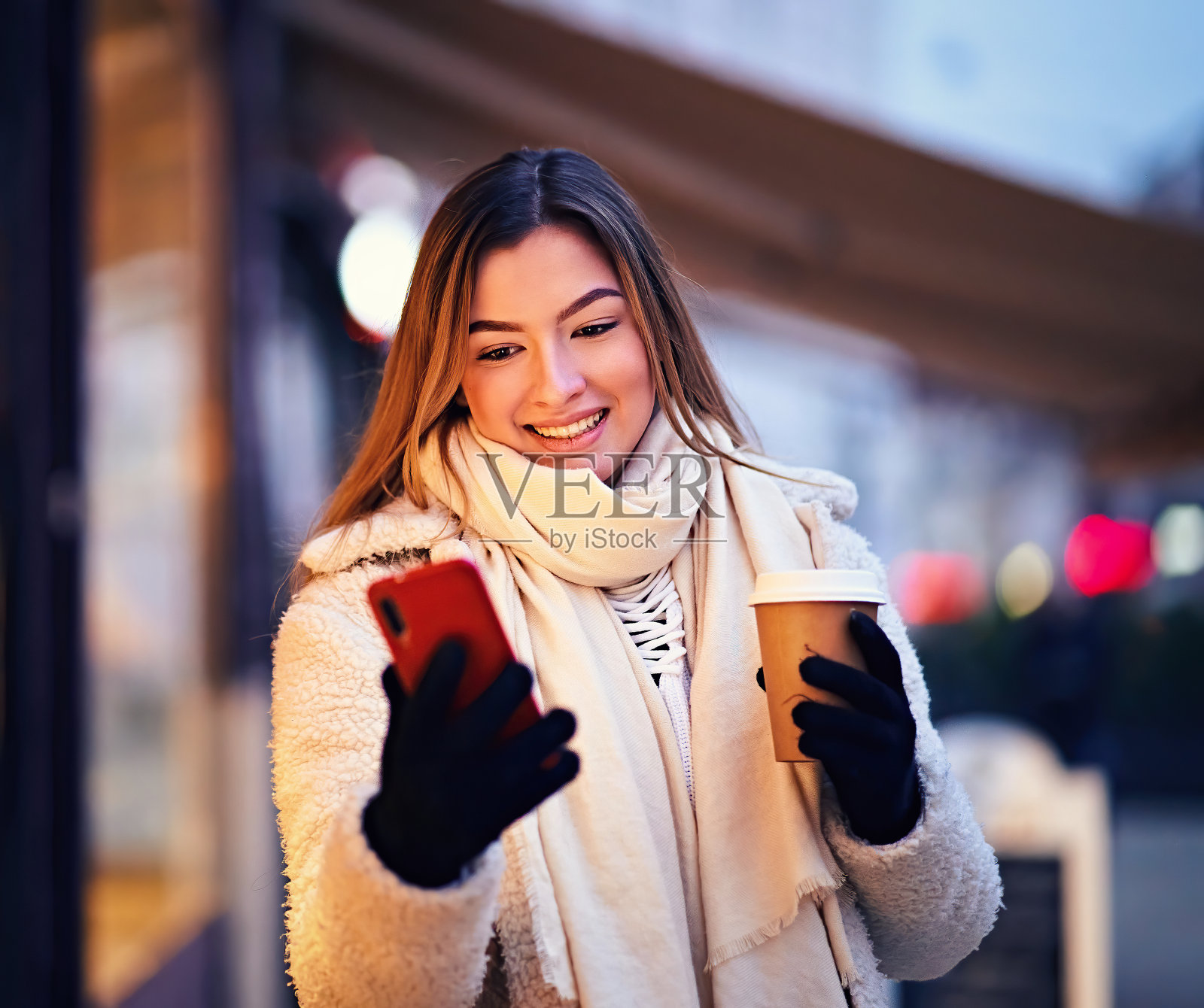 年轻快乐微笑的少女走在城市的街道上，在一个寒冷的冬天的晚上，夹克和外卖咖啡在她的手，同时浏览她的手机上的互联网照片摄影图片