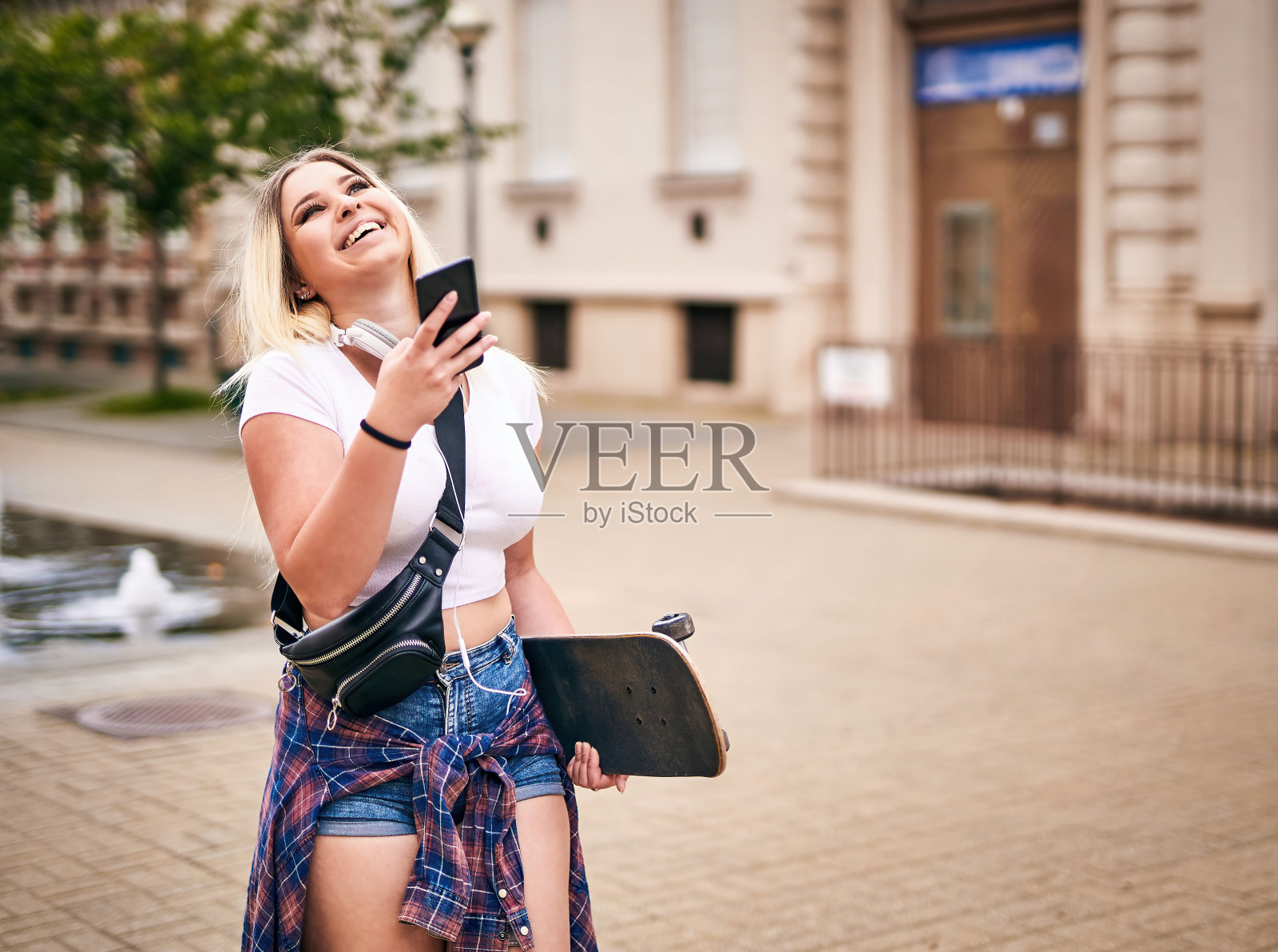 欢快的千禧一代女学生手拿滑板走在街上，用智能手机分享故事——现代数字通信概念照片摄影图片