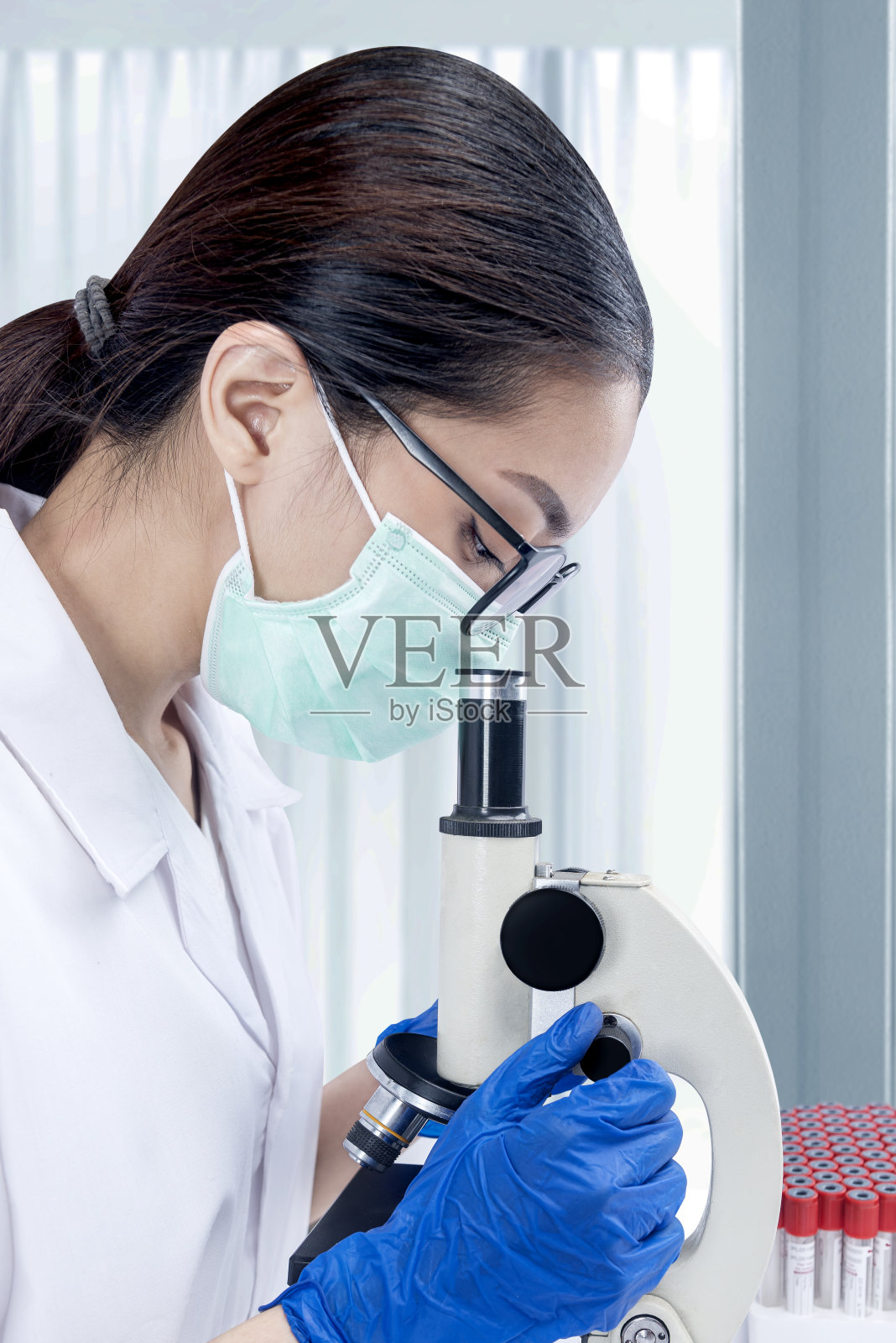 戴着面罩和眼镜，用显微镜观察的亚洲女研究员照片摄影图片