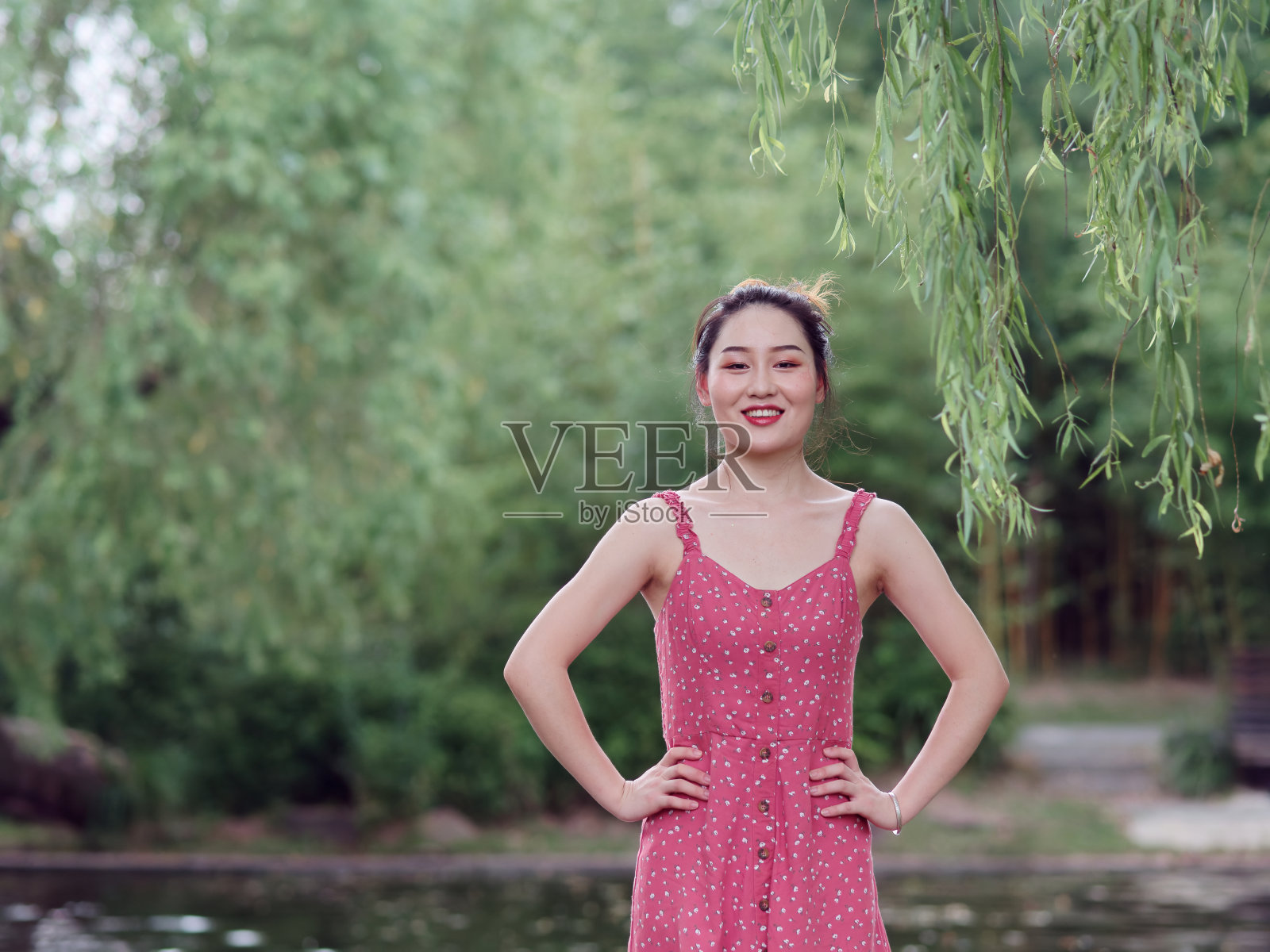 年轻的亚洲妇女的肖像看着和微笑与柳树的树枝在森林公园，美丽的中国女孩在性感的红色连衣裙享受她的无忧无虑的时间在炎热的夏天照片摄影图片