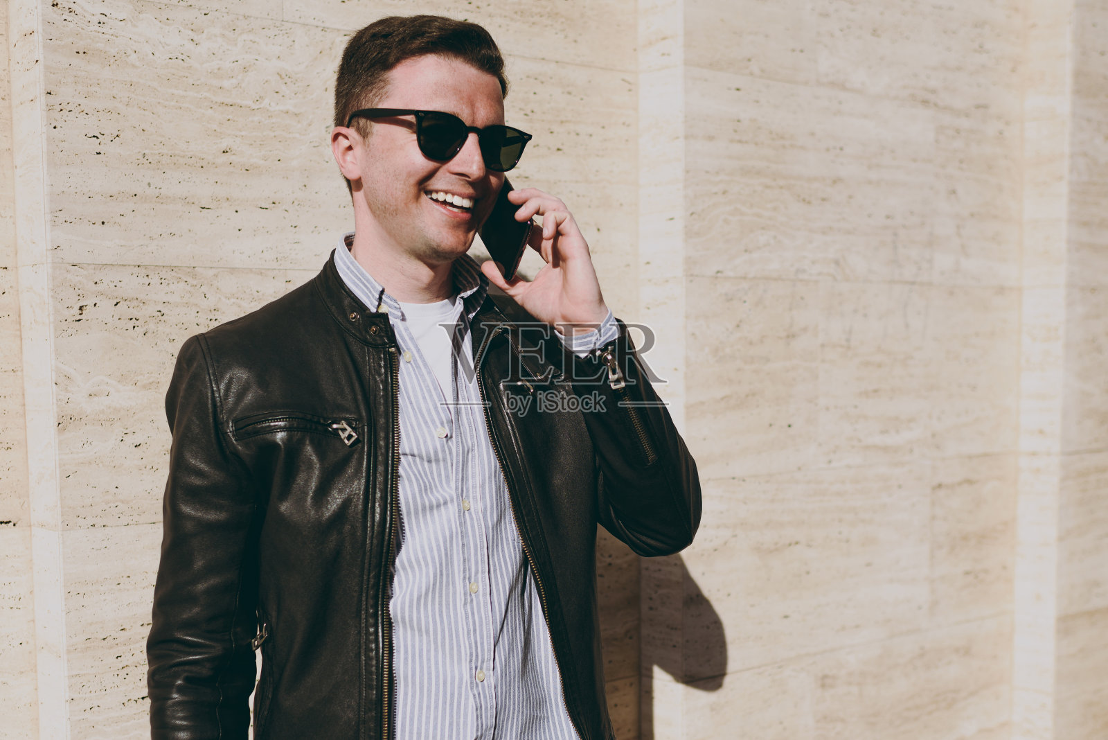 年轻的快乐快乐的男子穿着皮夹克衬衫眼镜站在户外的建筑墙壁背景上，用手机交谈，摊开双手旁观人们的城市生活方式和服装概念照片摄影图片