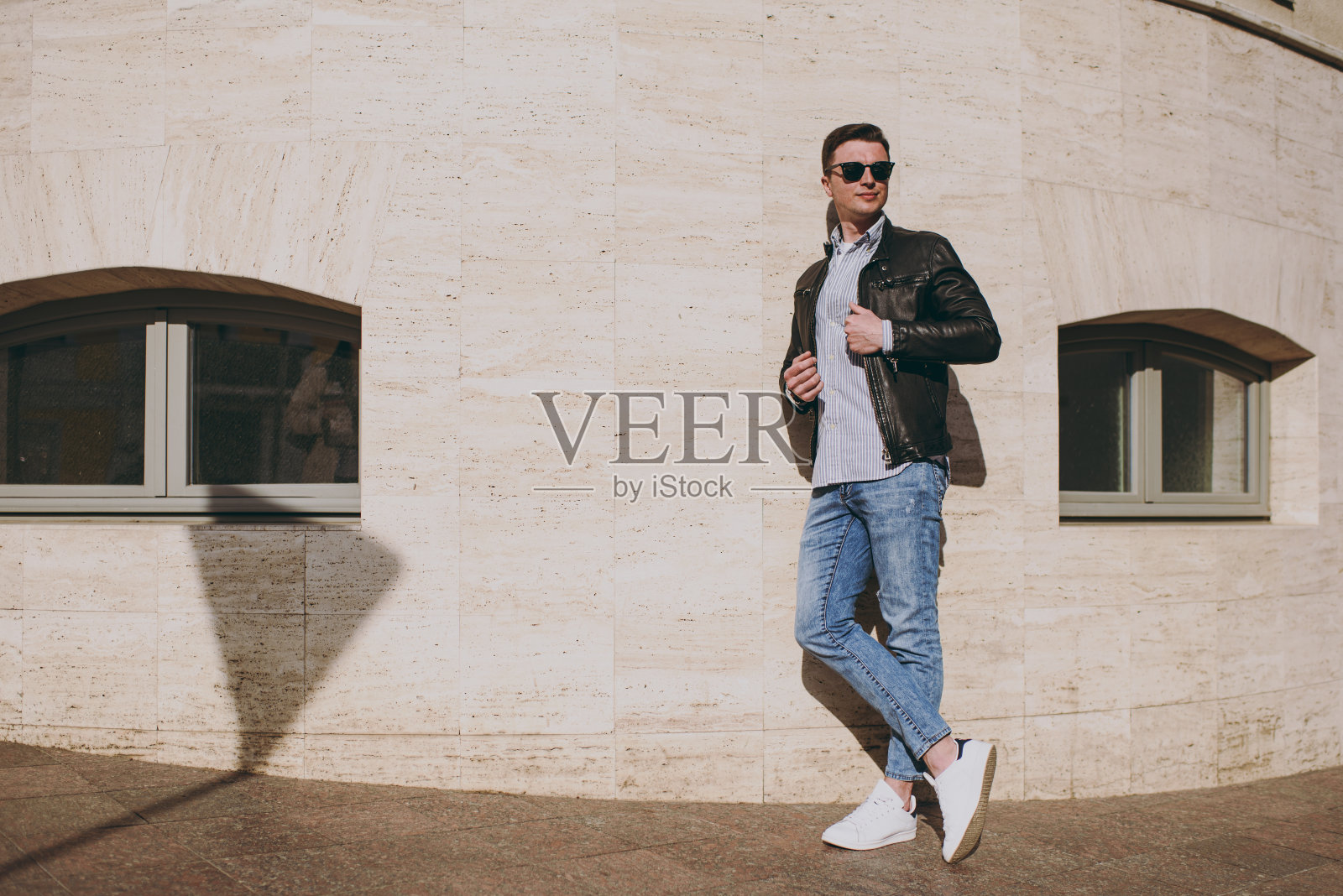 年轻的全长时尚微笑时尚快乐的高加索时尚男子，穿着皮夹克衬衫眼镜站在户外建筑墙旁，望向一边的概念，人们的城市生活方式和服装照片摄影图片