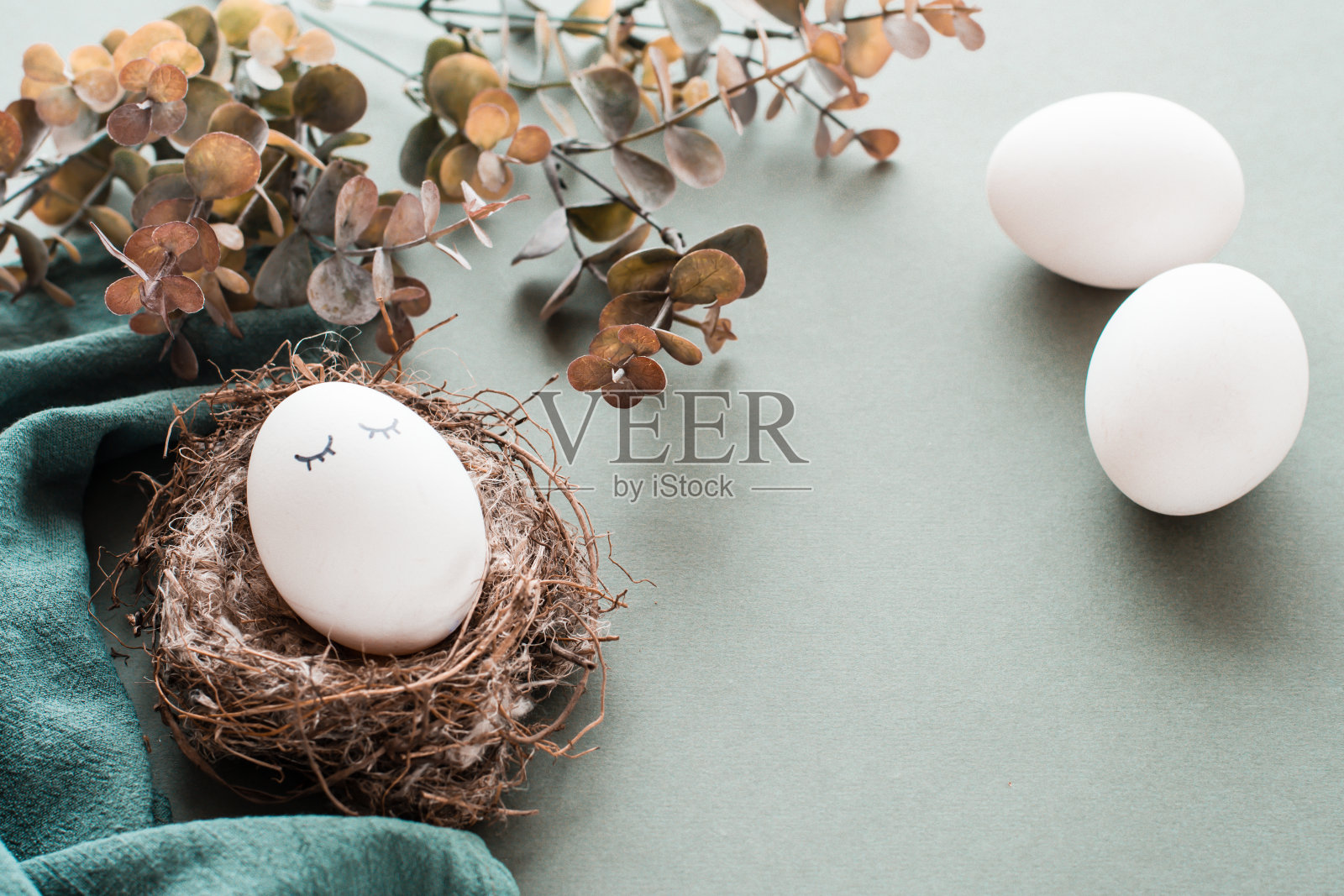 白色的鸡蛋与眼睛在巢，亚麻餐巾，其他鸡蛋和桉树树枝上的绿色背景。环保复活节快乐。照片摄影图片