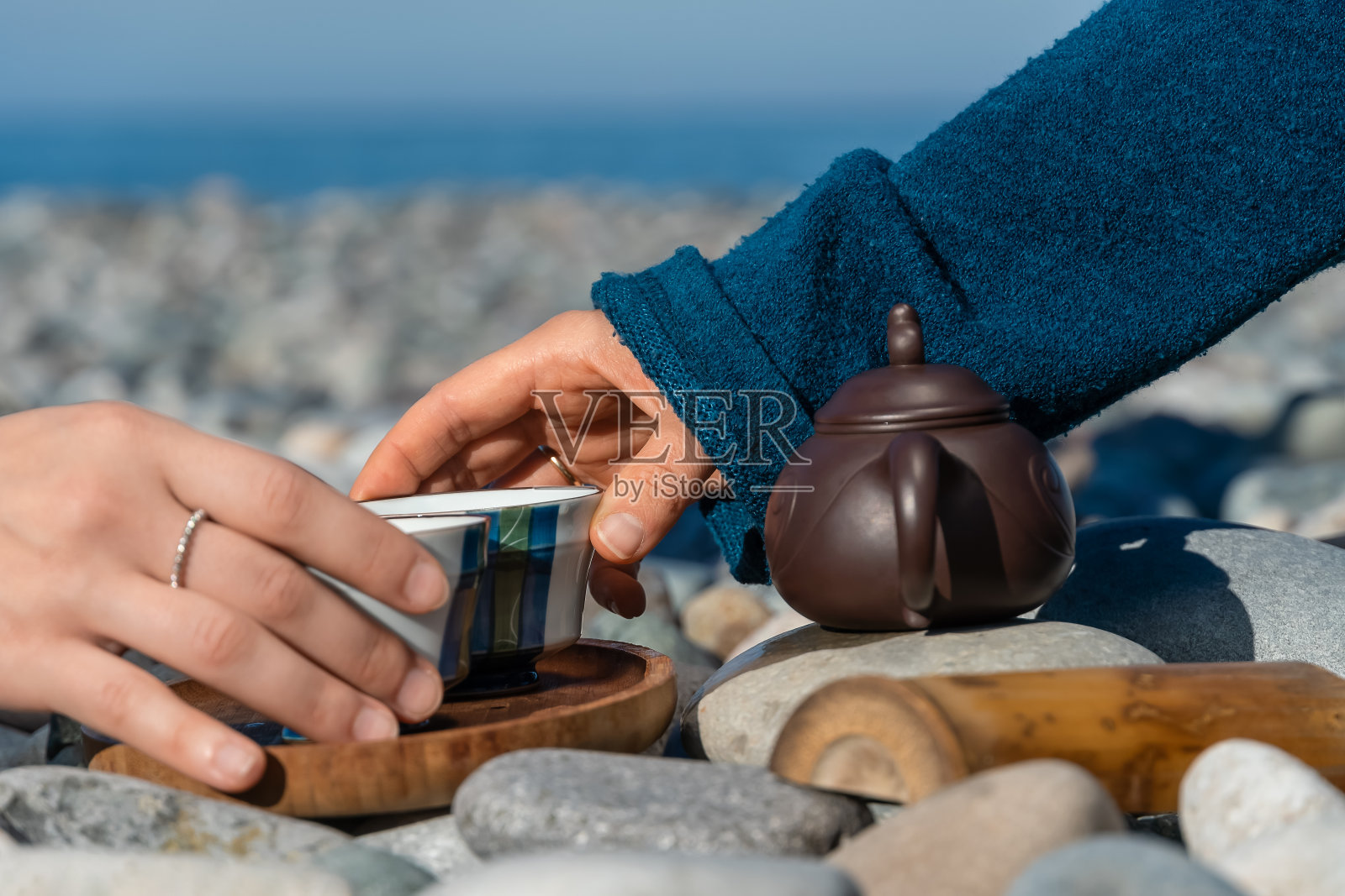 特写镜头，两个女人的手从一个木托盘上拿着茶杯，旁边有一个特别的茶壶。阳光明媚的一天，卵石海滩上的茶道照片摄影图片