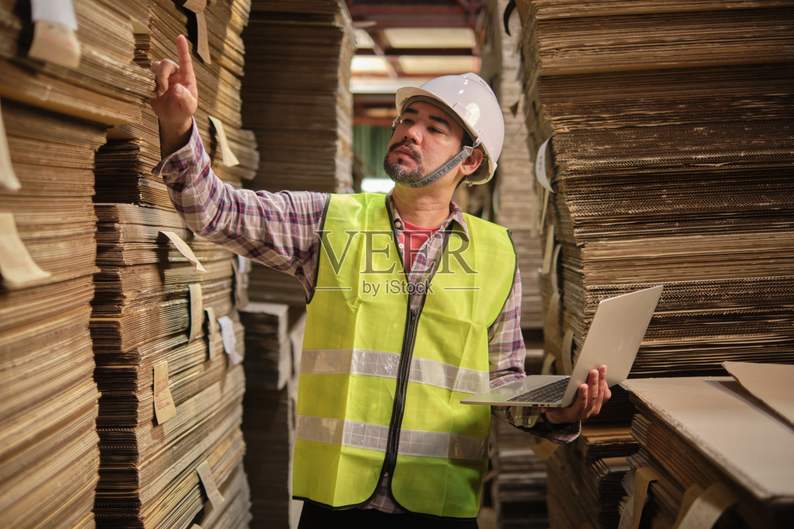 穿着制服的亚裔男性工人拿着笔记本电脑在纸厂仓库检查。照片摄影图片
