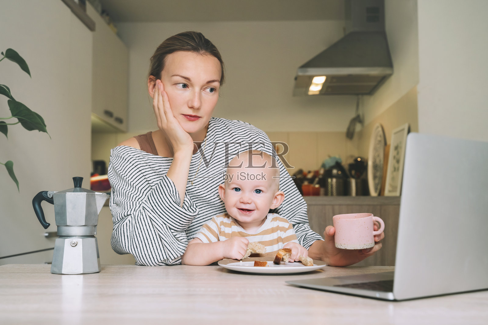 年轻的妈妈喝着咖啡，在厨房里和宝宝一起吃早餐。自由职业者，一夜未眠后的妈妈和孩子。休产假在家在线学习或工作的妇女照片摄影图片