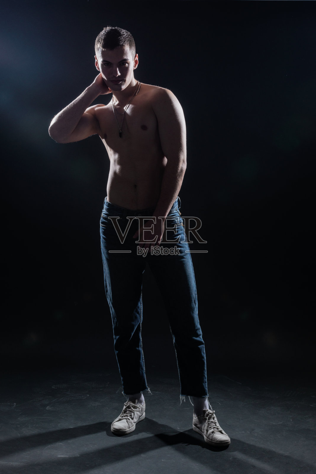 一个上身赤裸的肌肉男穿着牛仔裤在黑暗的摄影棚里摆姿势的剪影。照片摄影图片