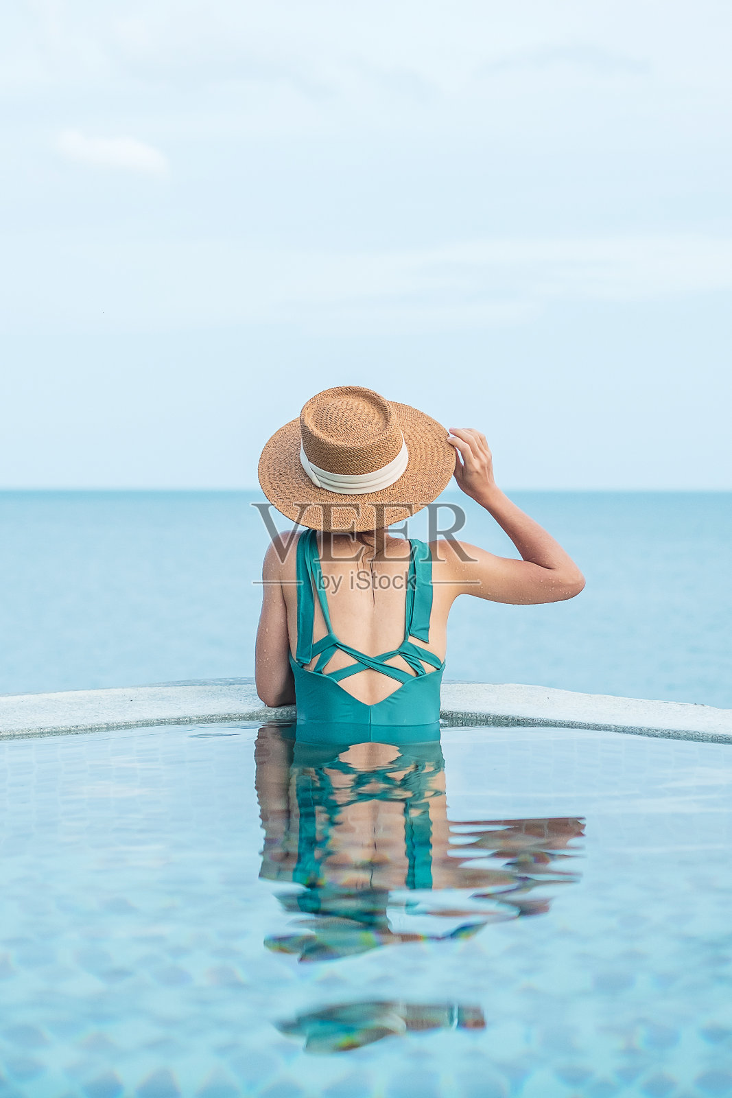 快乐的女人在绿色的泳衣和帽子在豪华酒店的游泳池对着海洋。年轻女性喜欢在热带度假胜地。放松，夏季，旅游，假期，假期和周末的概念照片摄影图片