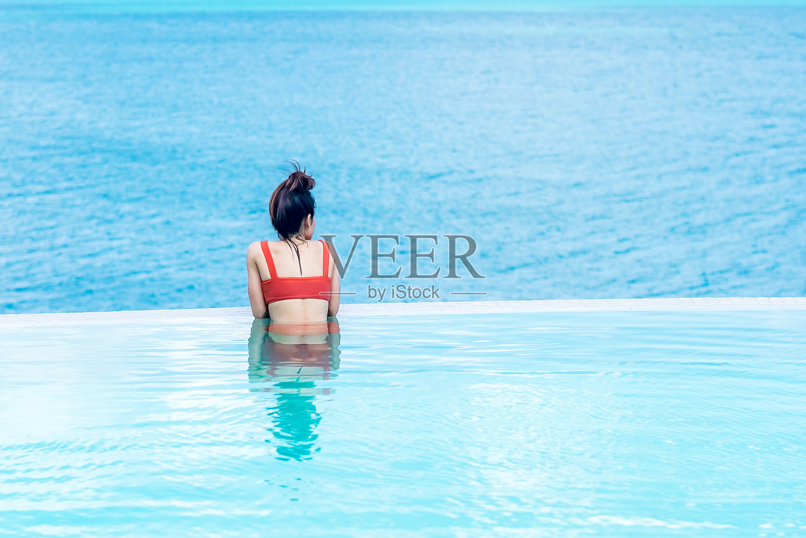 快乐的女人在橙色泳衣游泳在豪华酒店的无边泳池对着海洋。年轻女性喜欢在热带度假胜地。放松，夏季，旅游，假期，假期和周末的概念照片摄影图片