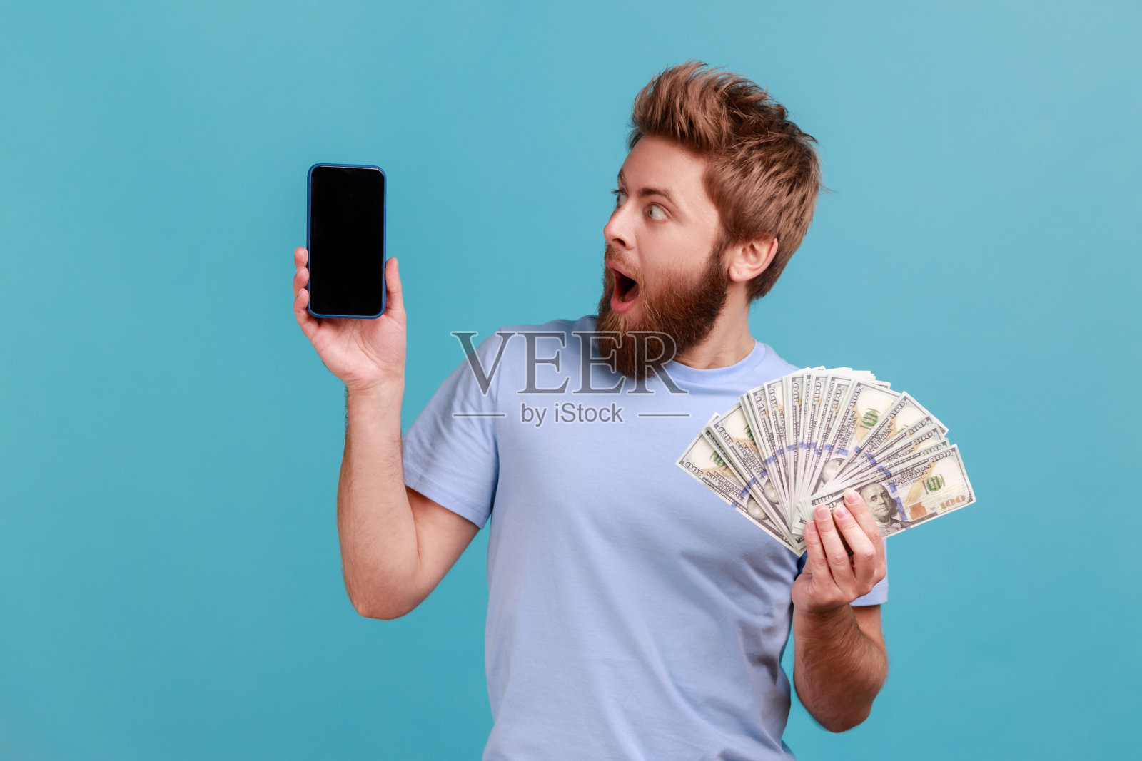 身穿蓝色t恤的男子拿着1美元钞票，看着屏幕空着的手机。照片摄影图片