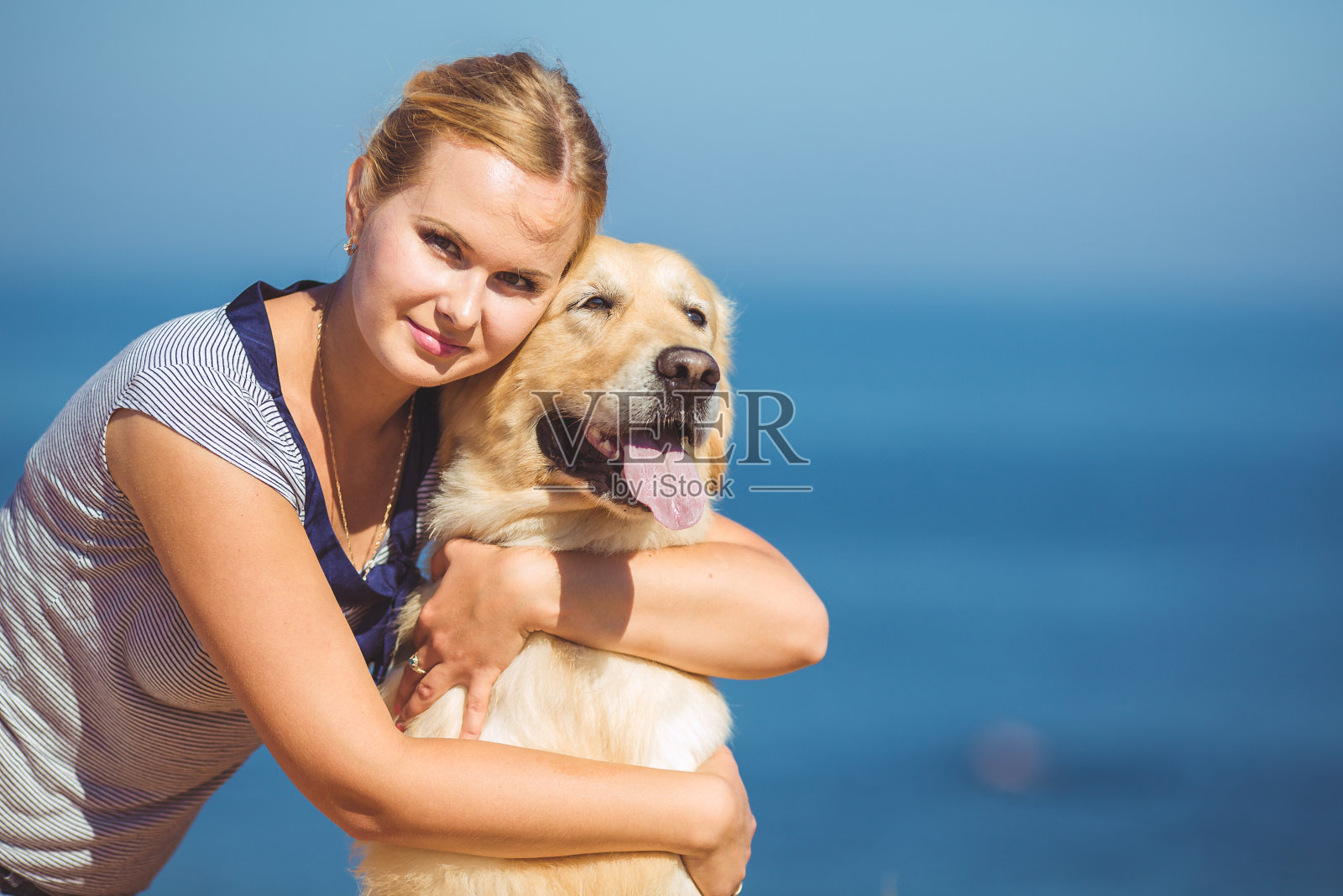 一个年轻的金发女人在蓝色海洋附近的夏日沙滩上训练她的拉布拉多狗朋友照片摄影图片