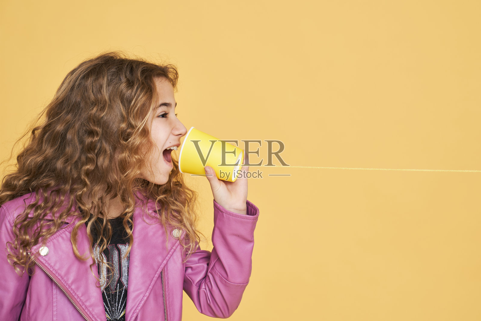 女孩透过一个纸杯说话，纸杯绑在一个黄色背景的线孤立。照片摄影图片